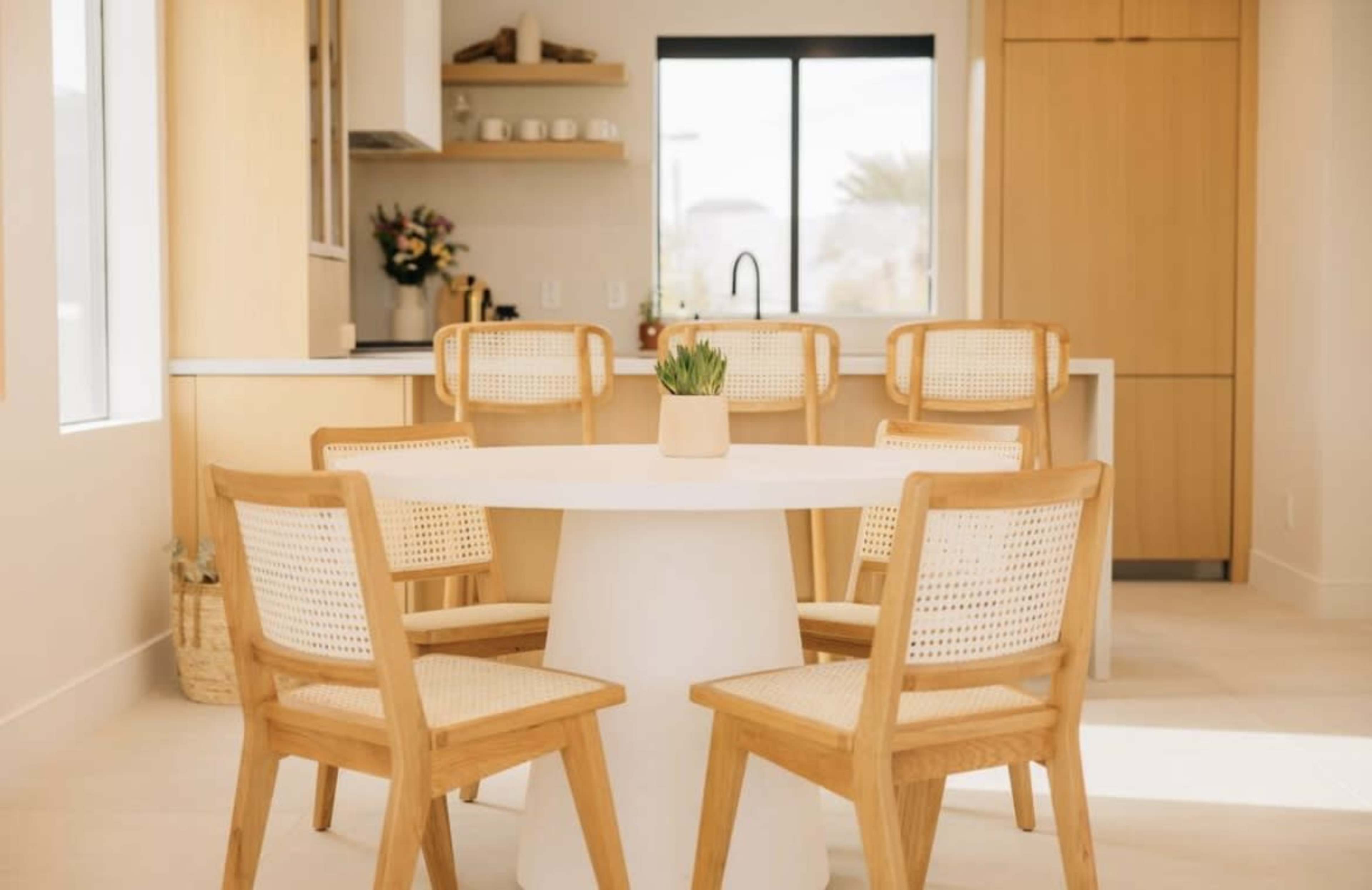 A round white dining table is surrounded by five wooden chairs with woven seats in a bright, modern kitchen space.