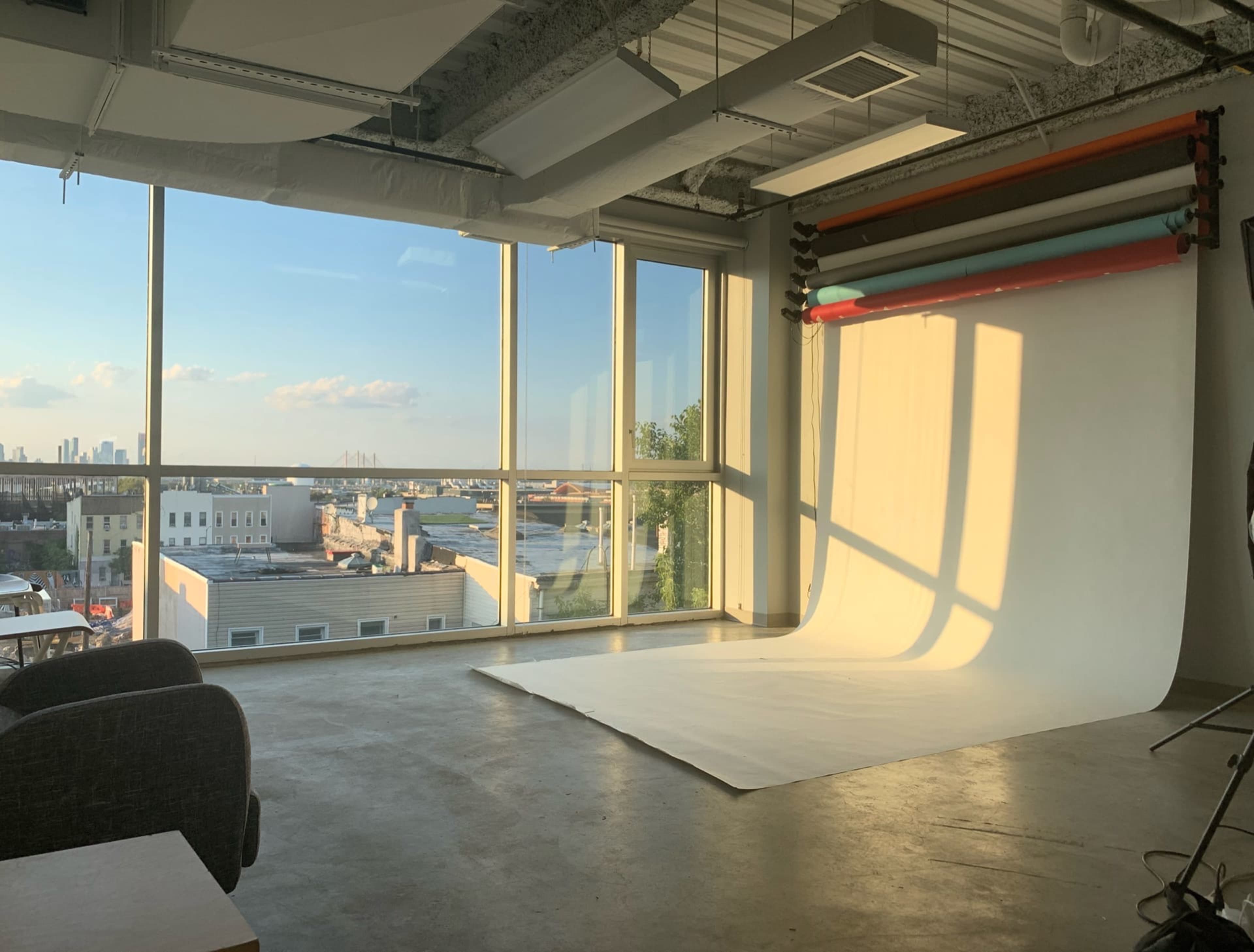 A photography studio with a large window overlooks a cityscape, featuring a white backdrop and colored rolls hanging on the wall.