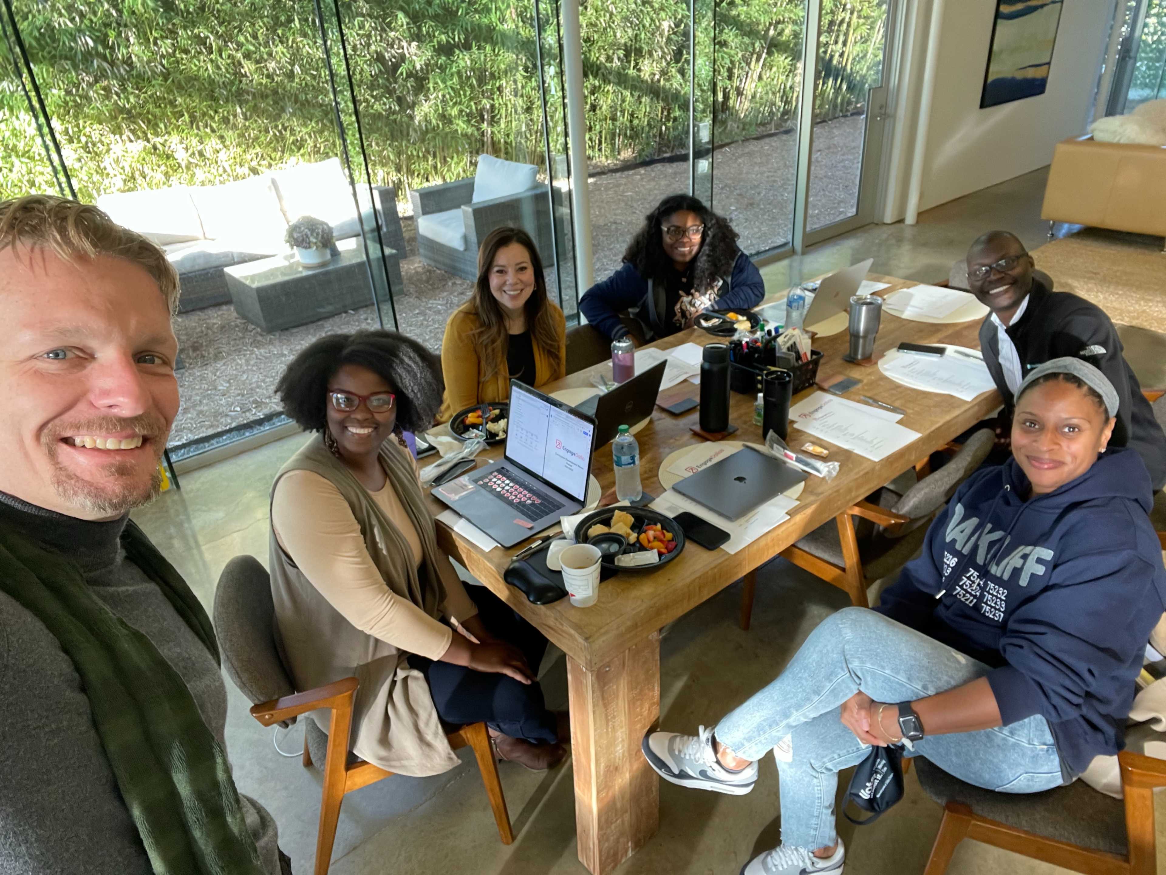A group of six people is gathered around a wooden table in a bright room with large glass windows, sharing food and work materials.