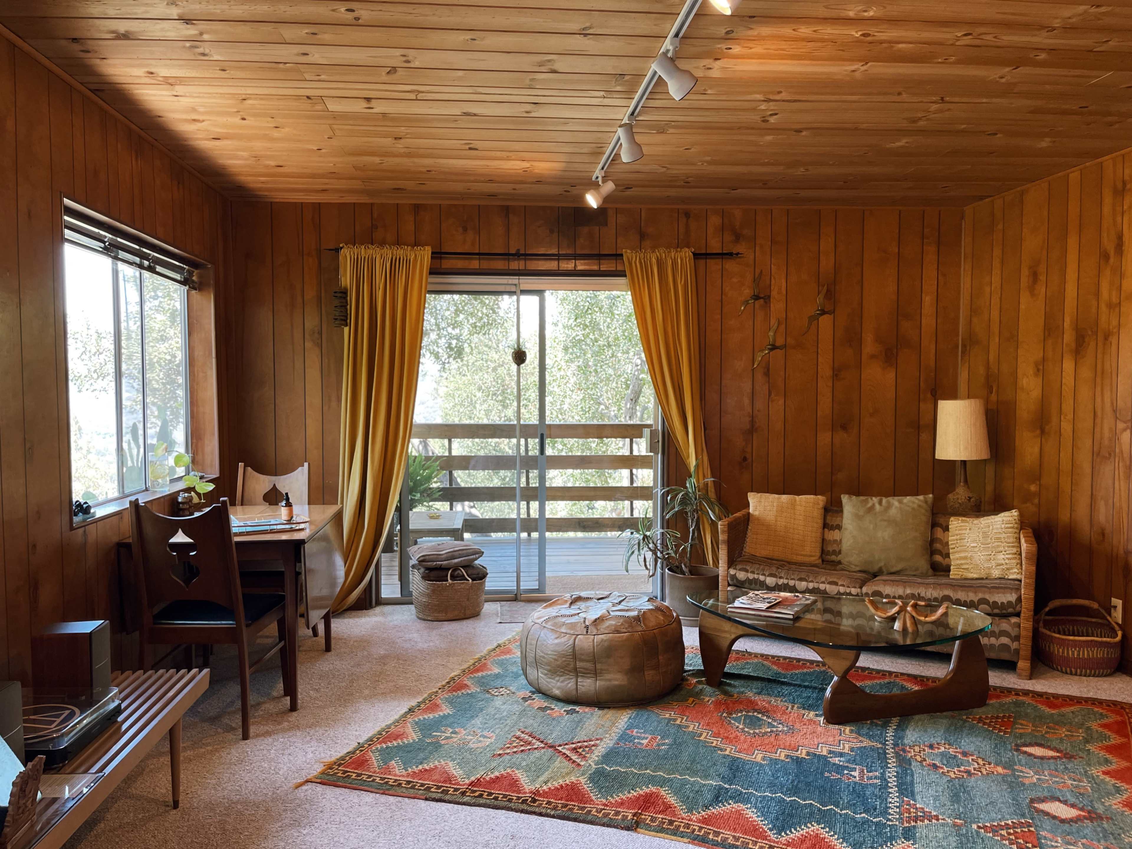 A cozy living room with wooden walls, a patterned rug, a sofa, and large windows leading to a view outside.