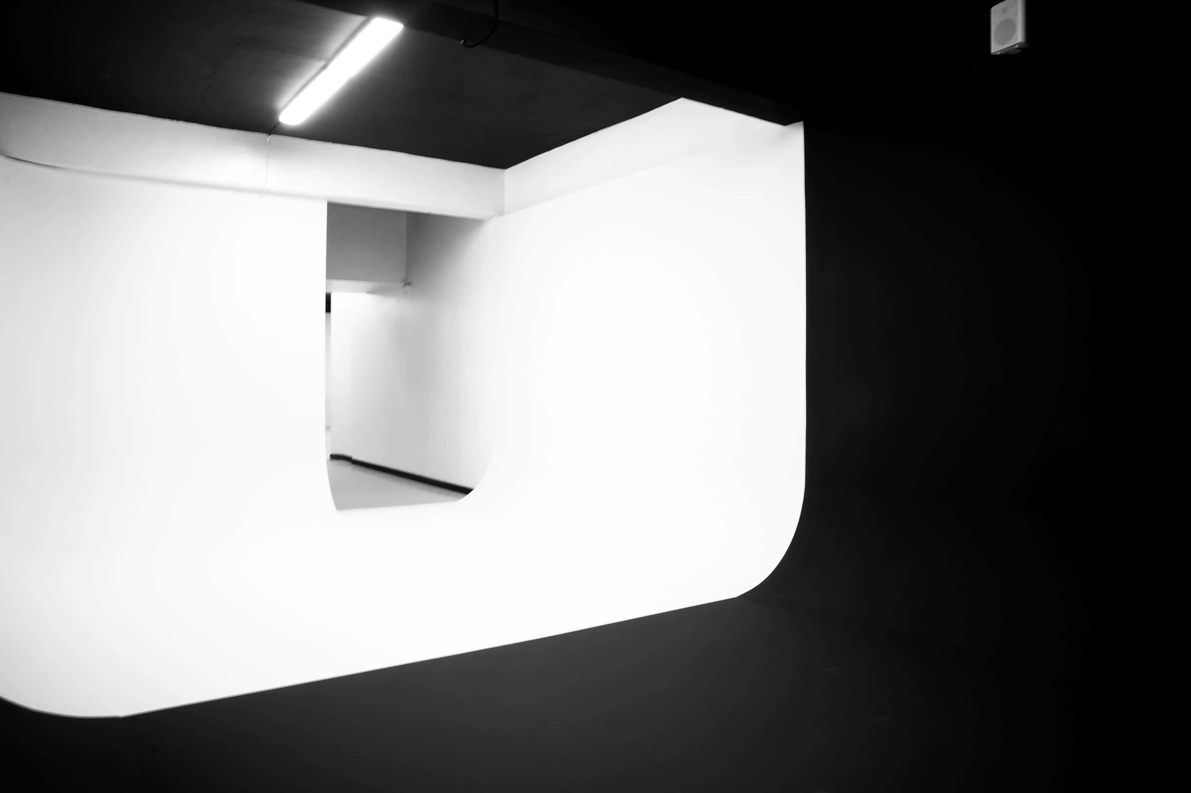The image depicts a minimalist interior space featuring a curved white wall against a black background, with a hallway visible in the distance.