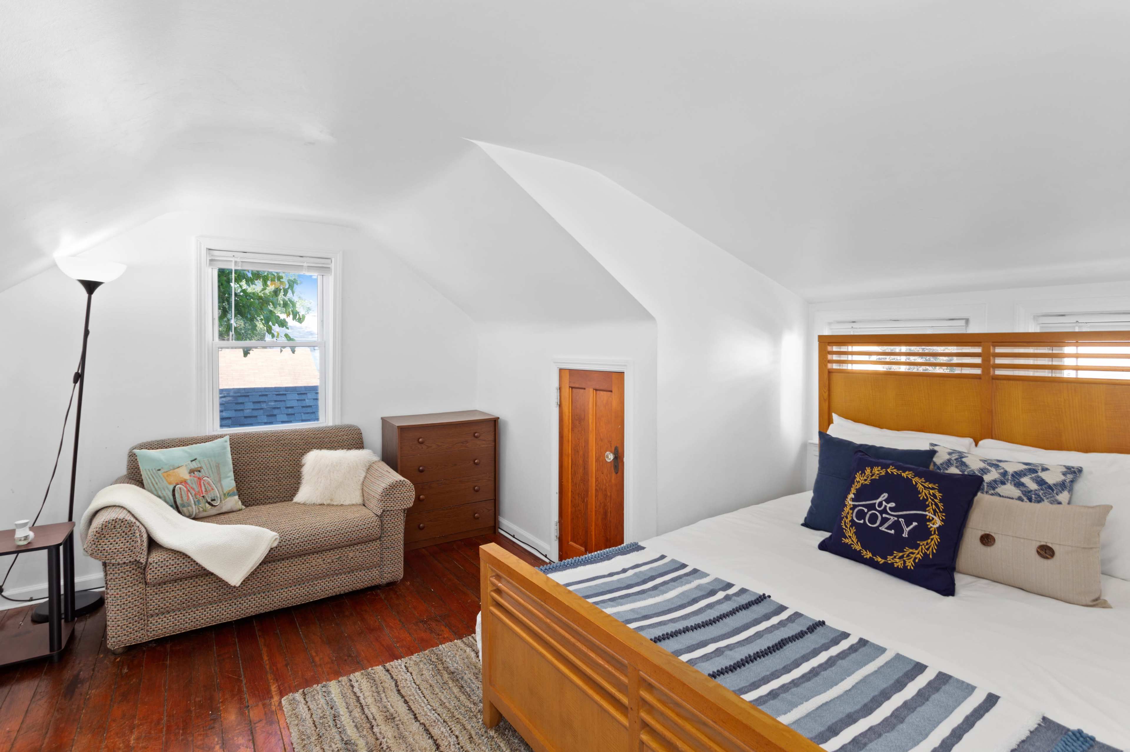 A well-lit bedroom features a wooden bed with decorative pillows, a cozy sofa, a chest of drawers, and a door, all set against white walls and a sloped ceiling.