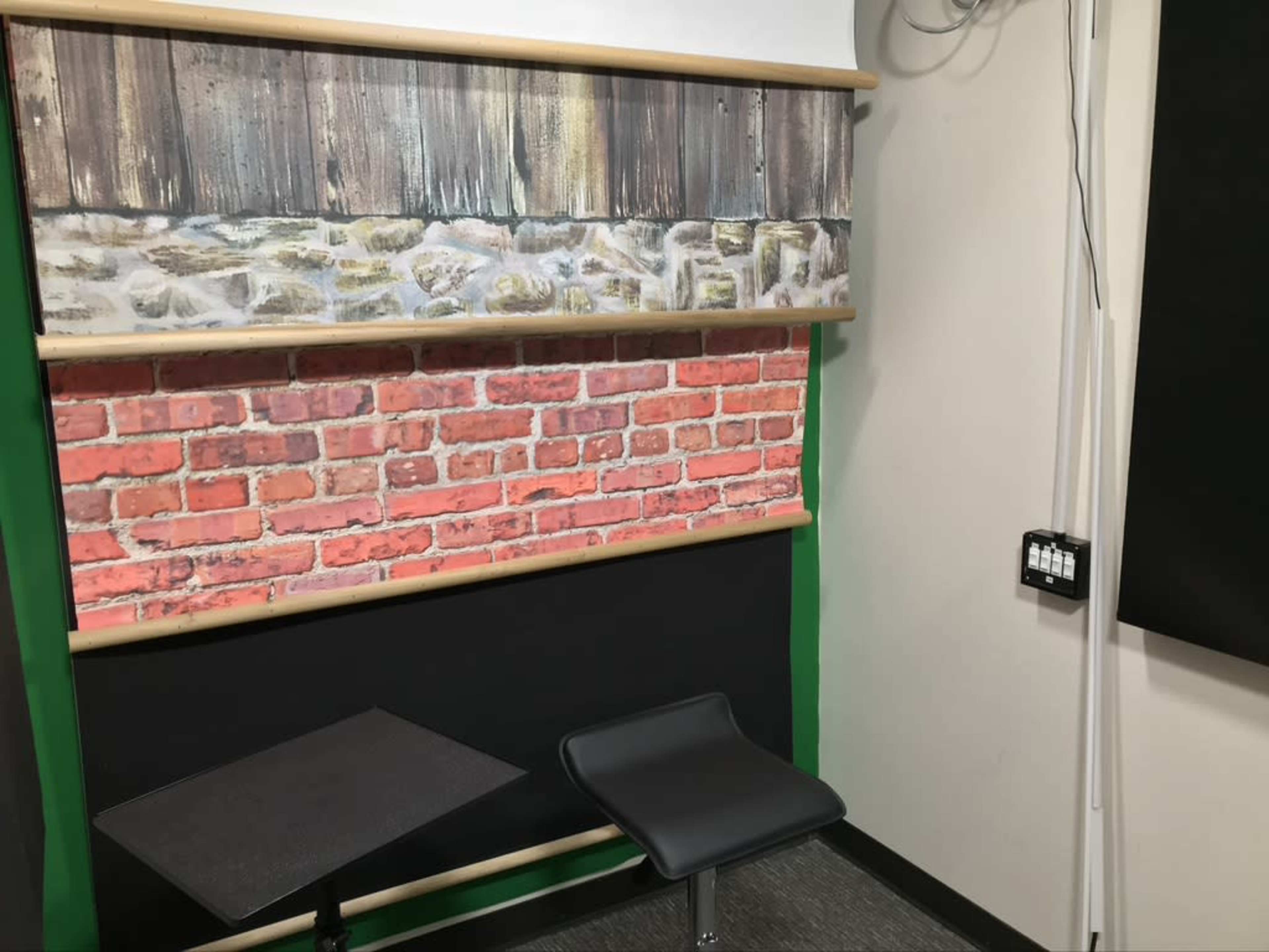 The image shows a corner of a room featuring a wall with a wood and stone pattern above a red brick wall, alongside a small black table and a single chair.