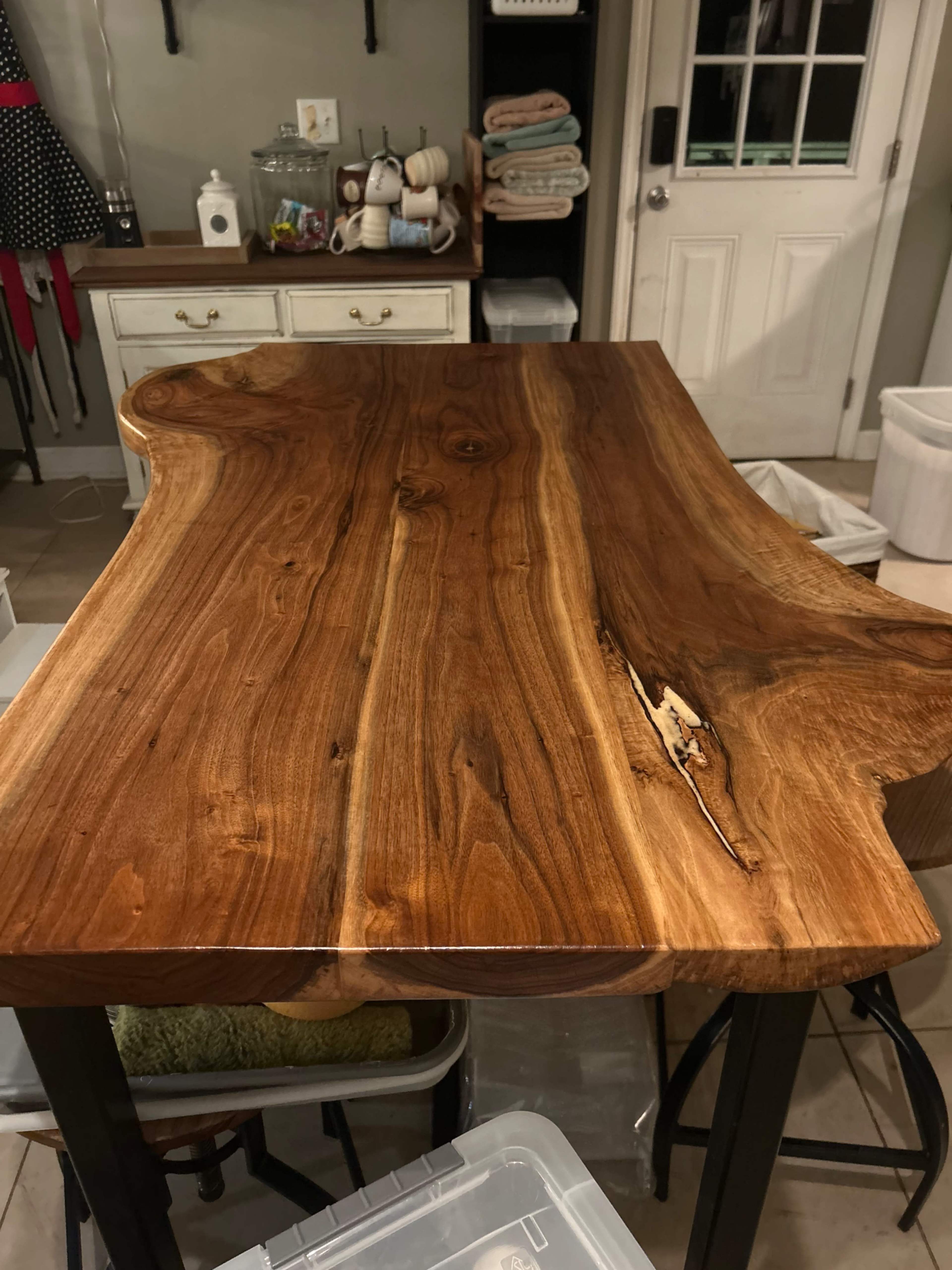 A wooden table with a natural edge design, featuring prominent grain patterns and a smooth finish, positioned in a room with a light-colored wall and storage items in the background.