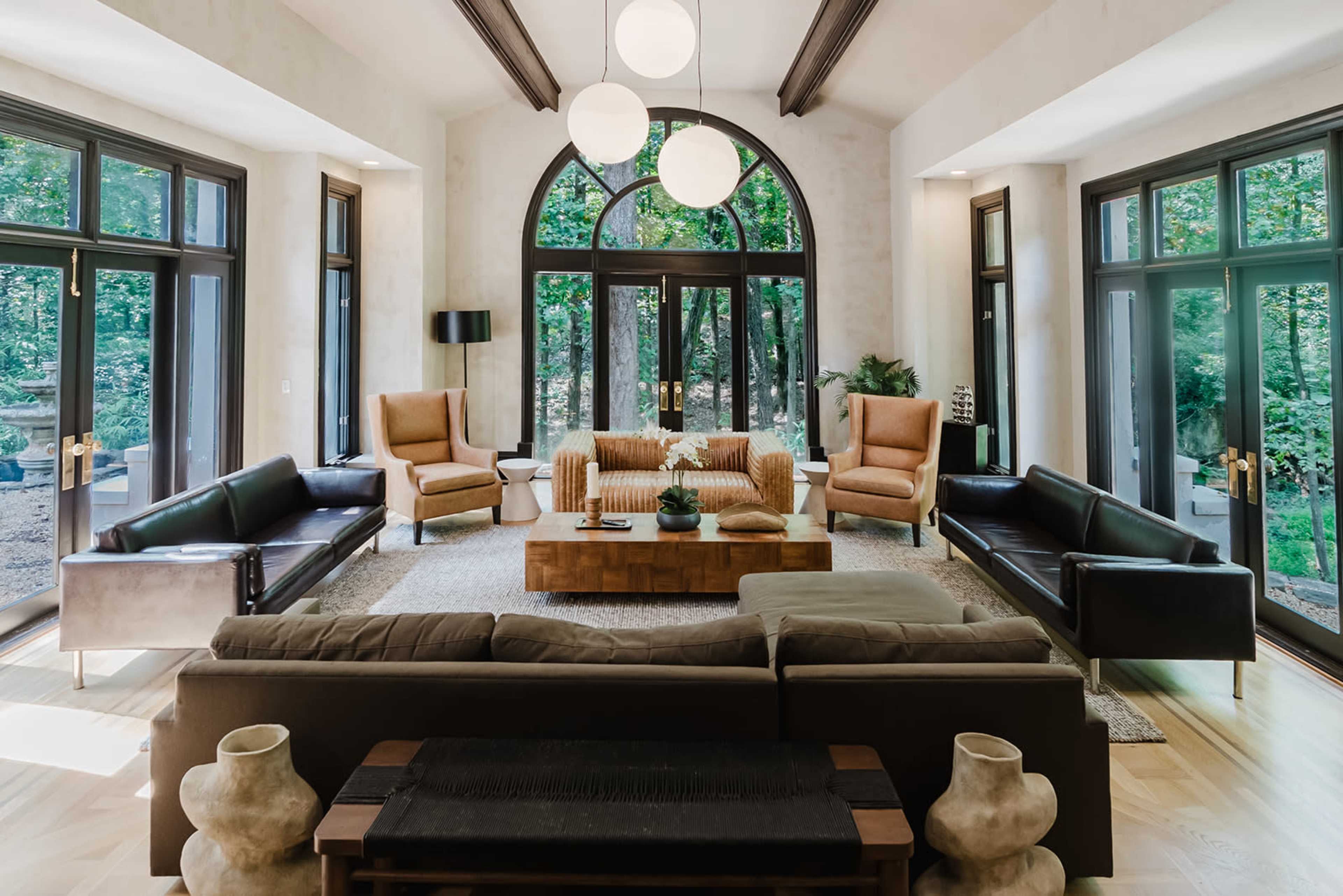 The image shows a spacious living room with large windows, featuring a mix of modern furniture, including several armchairs, a coffee table, and a sectional sofa, all arranged around a central area rug.