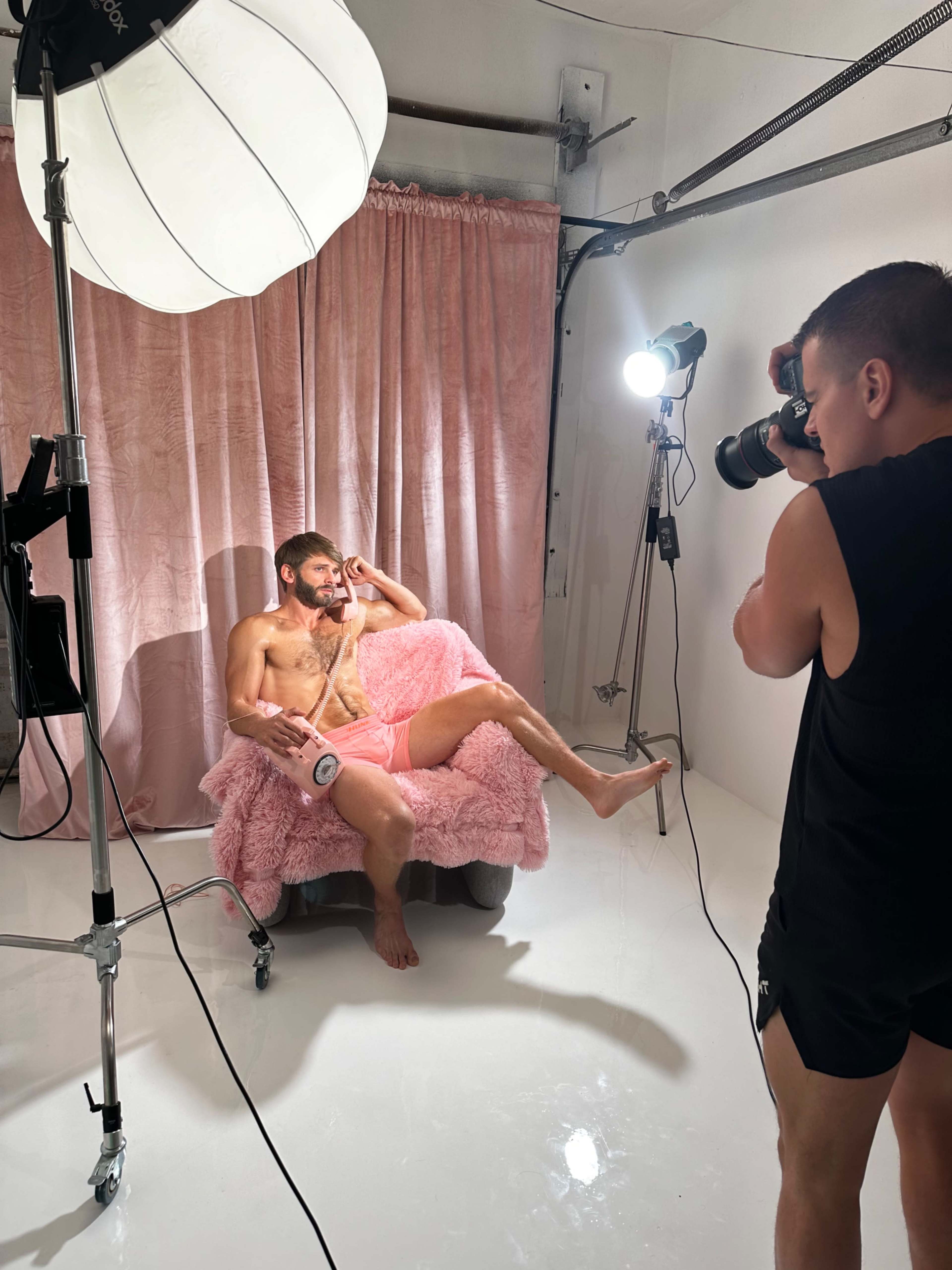 A man sits on a pink fuzzy couch while another man photographs him in a well-lit studio setting.