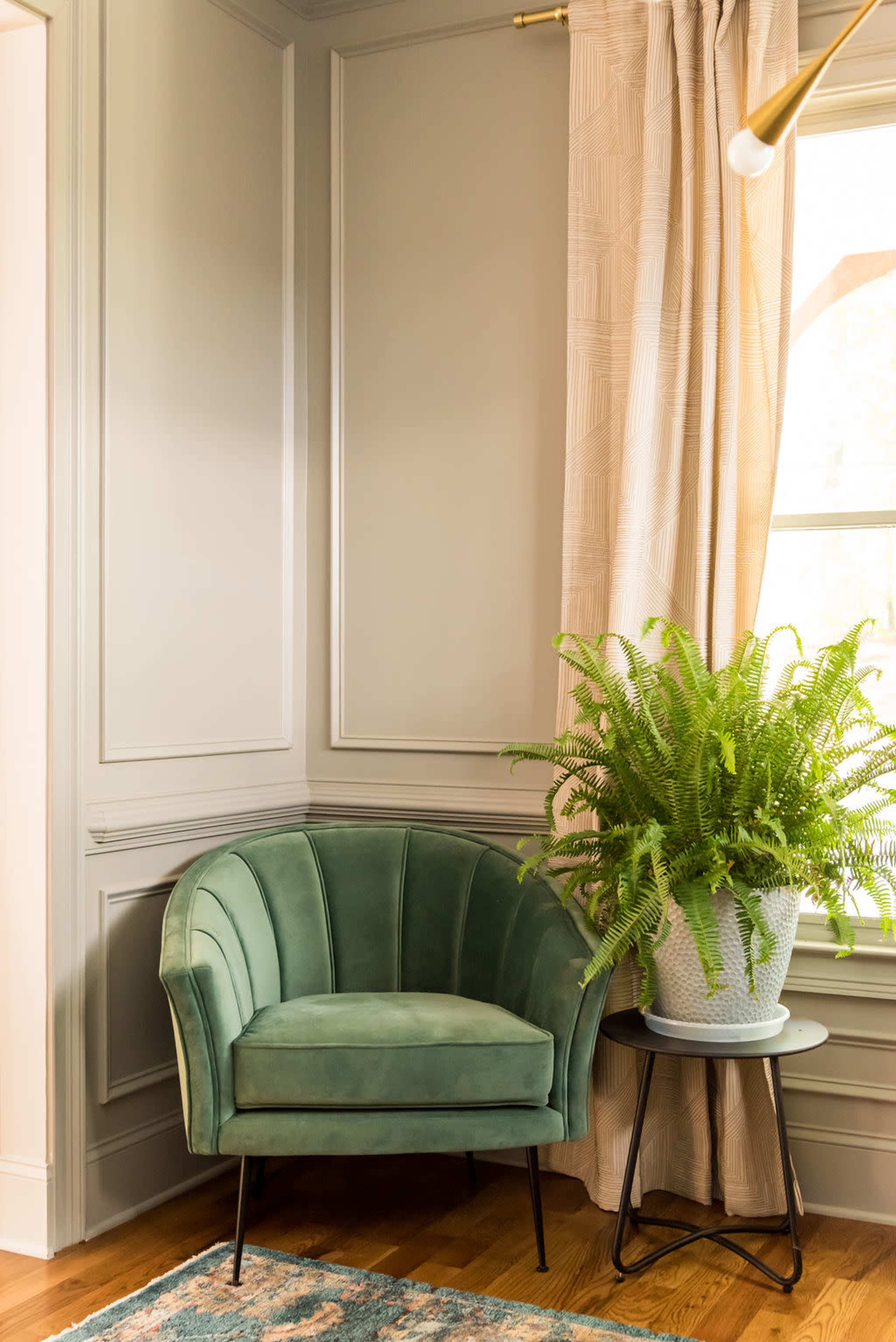 A green upholstered chair is positioned next to a tall potted fern and a side table in a corner of a room with light-colored walls.
