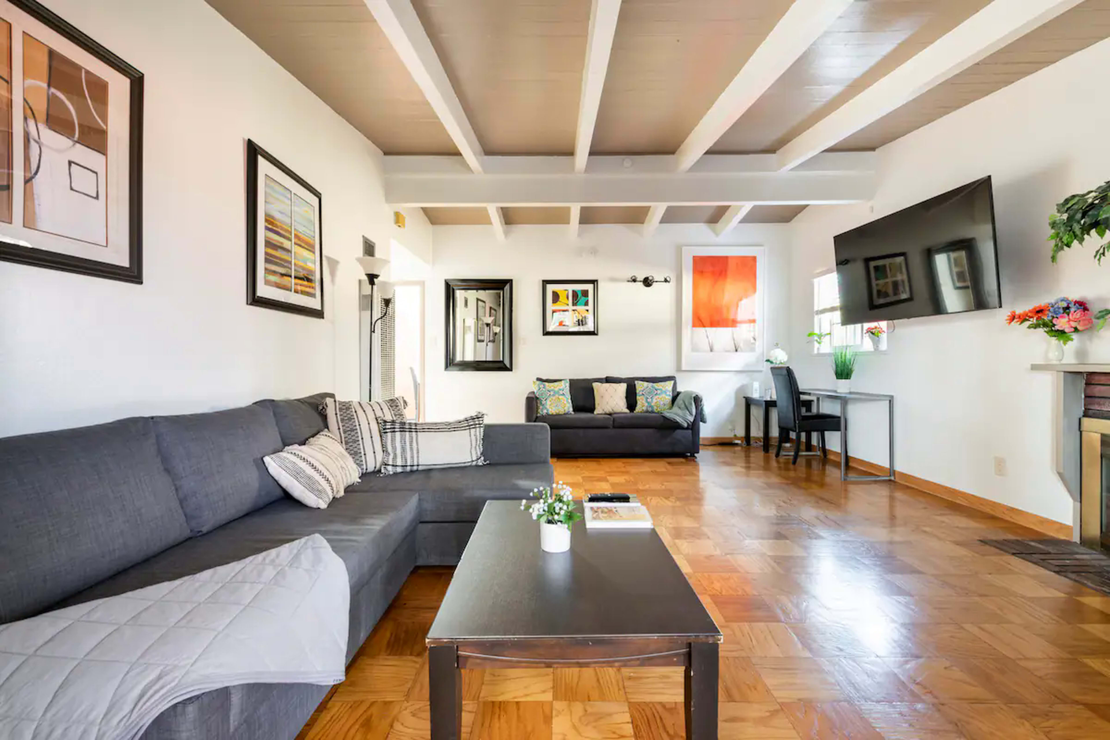 A living room with two sofas, a coffee table, a TV mounted on the wall, and a dining area in the background.
