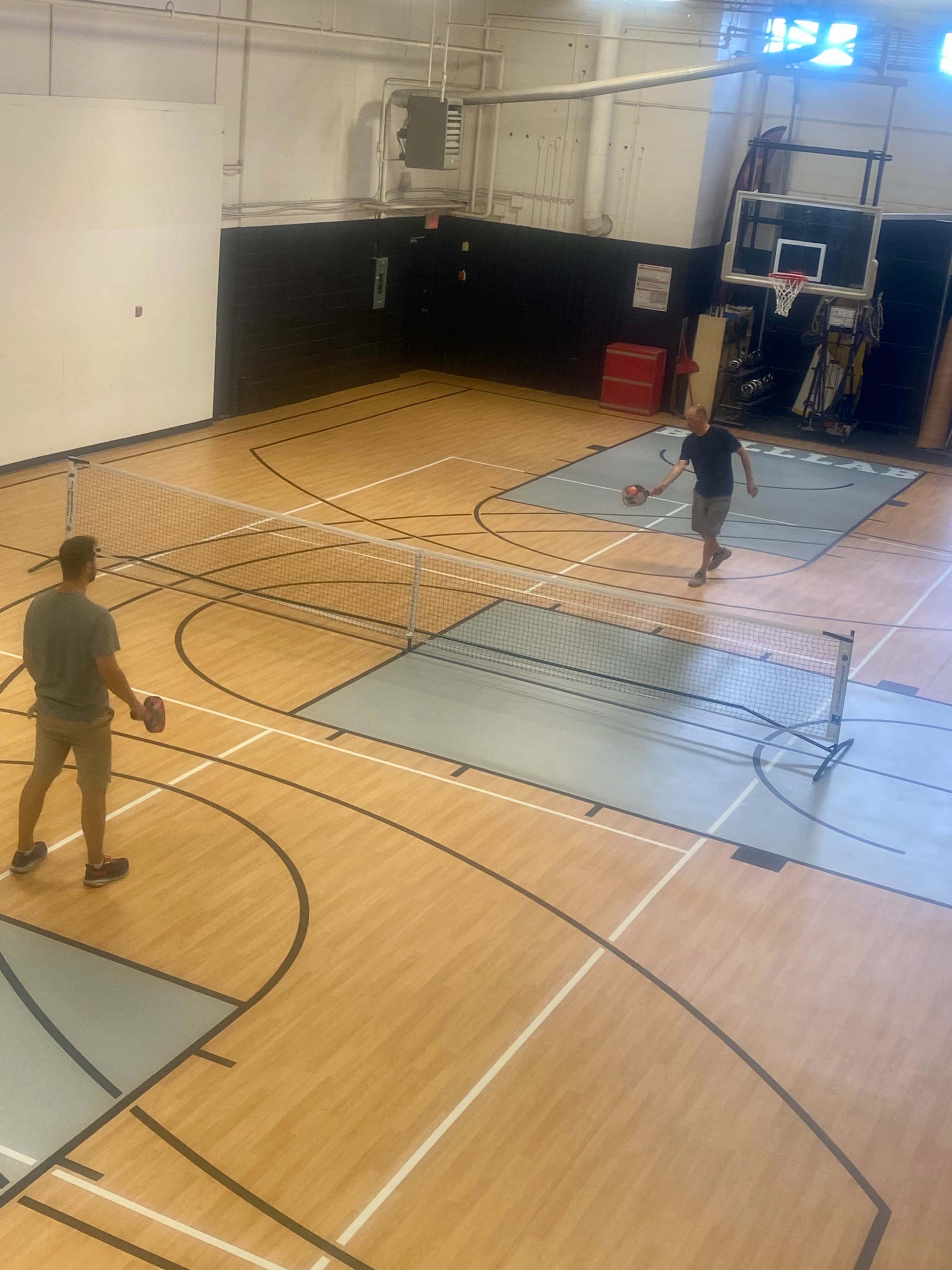 Two people are playing pickleball on a gymnasium court with nets set up in the center.