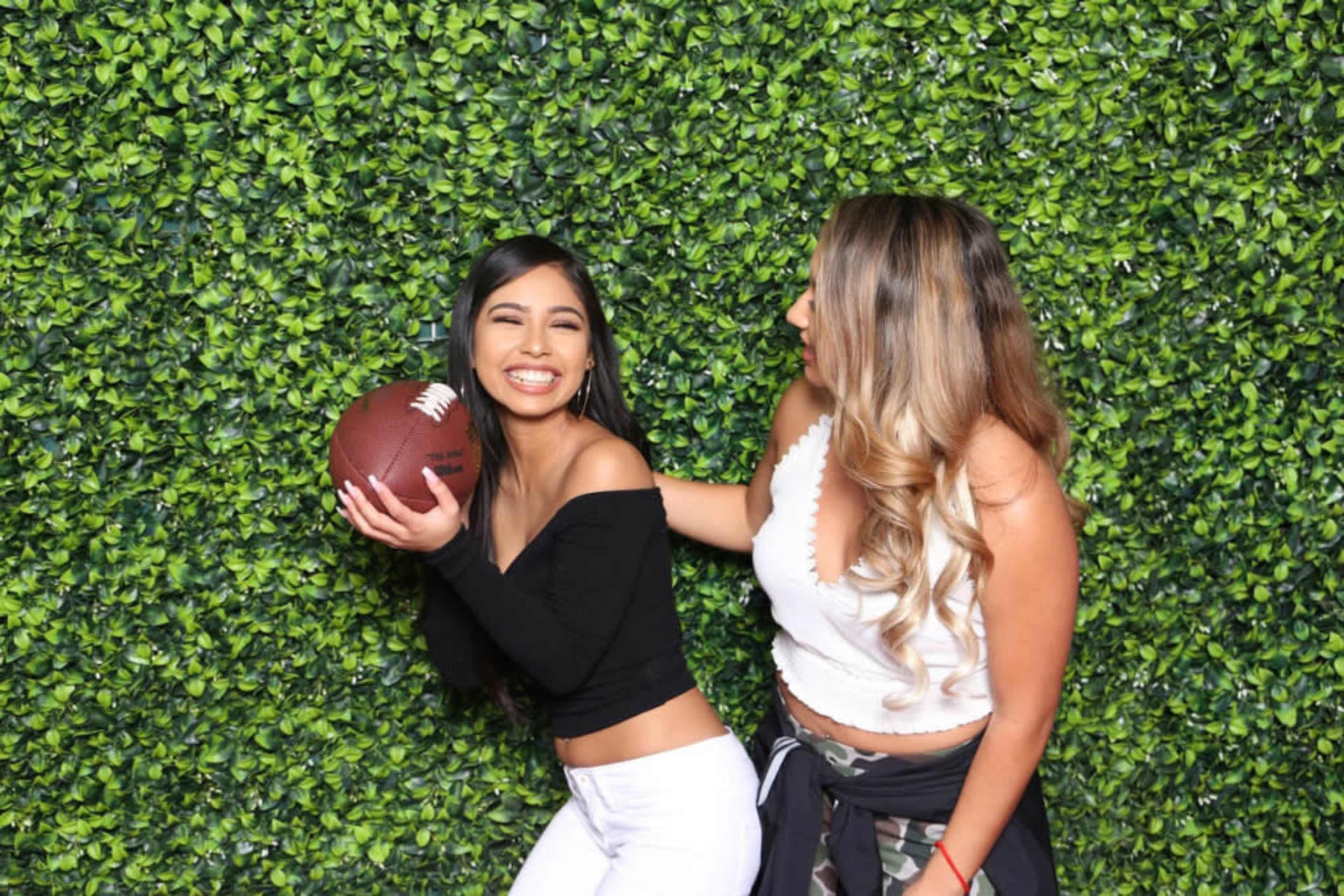 Two women pose together in front of a green leafy backdrop, with one holding a football and smiling while the other looks at her.