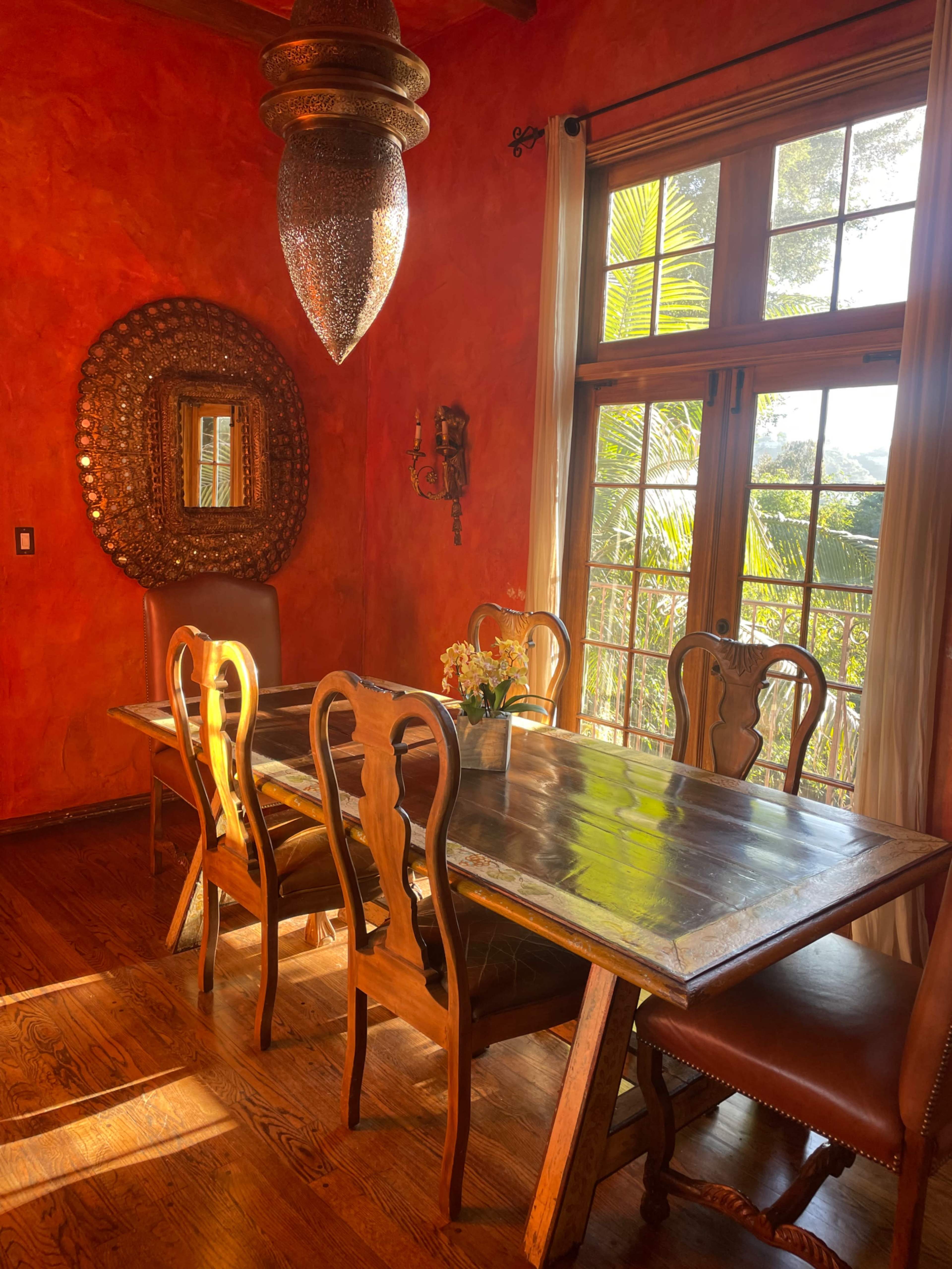 A wooden dining table surrounded by six chairs is illuminated by a hanging light fixture in a room with orange walls and large windows.