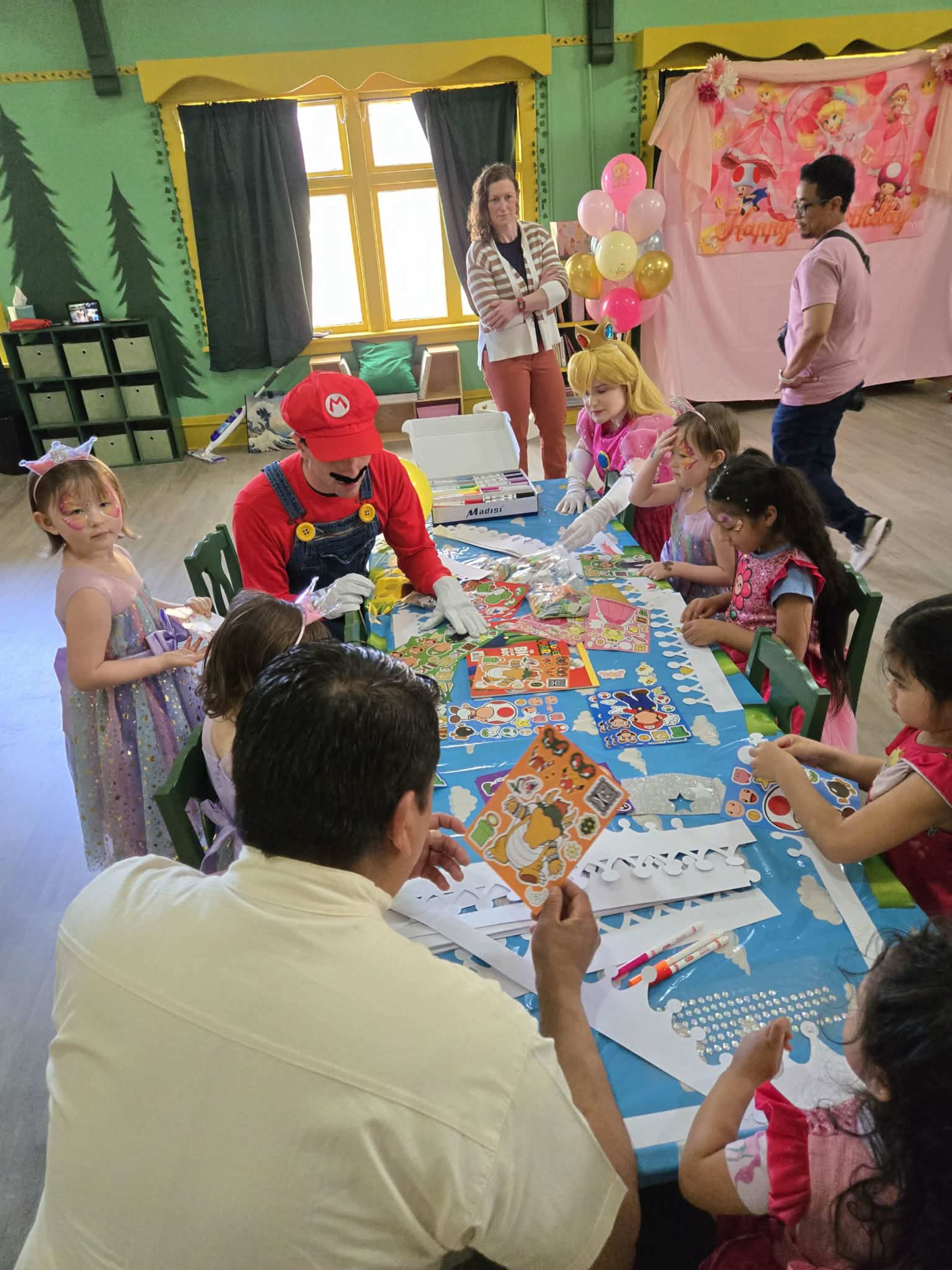 A group of children and adults are gathered around a table engaged in arts and crafts activities in a colorful party setting.