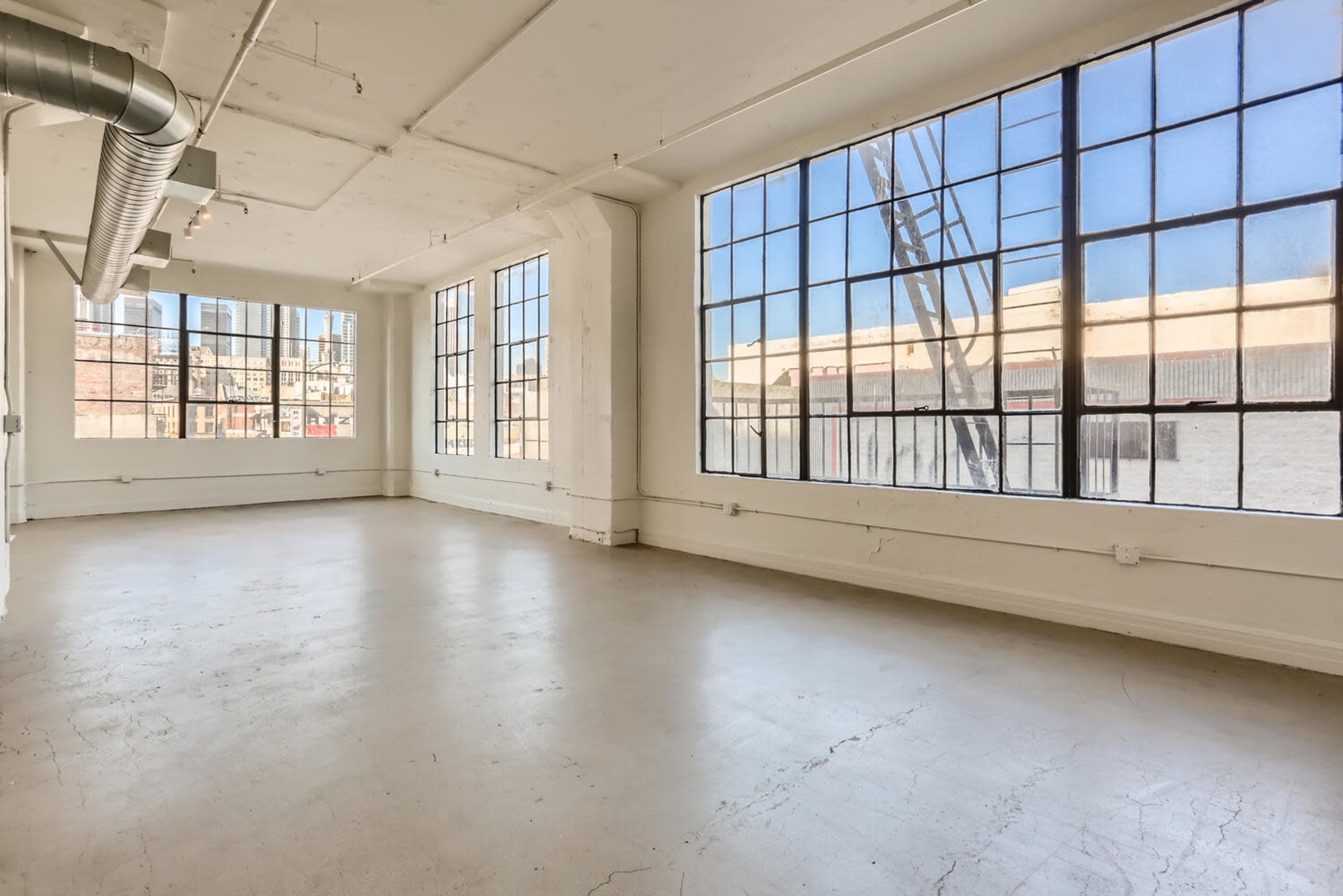 An empty room with large industrial windows and a concrete floor, featuring visible pipes along the ceiling.