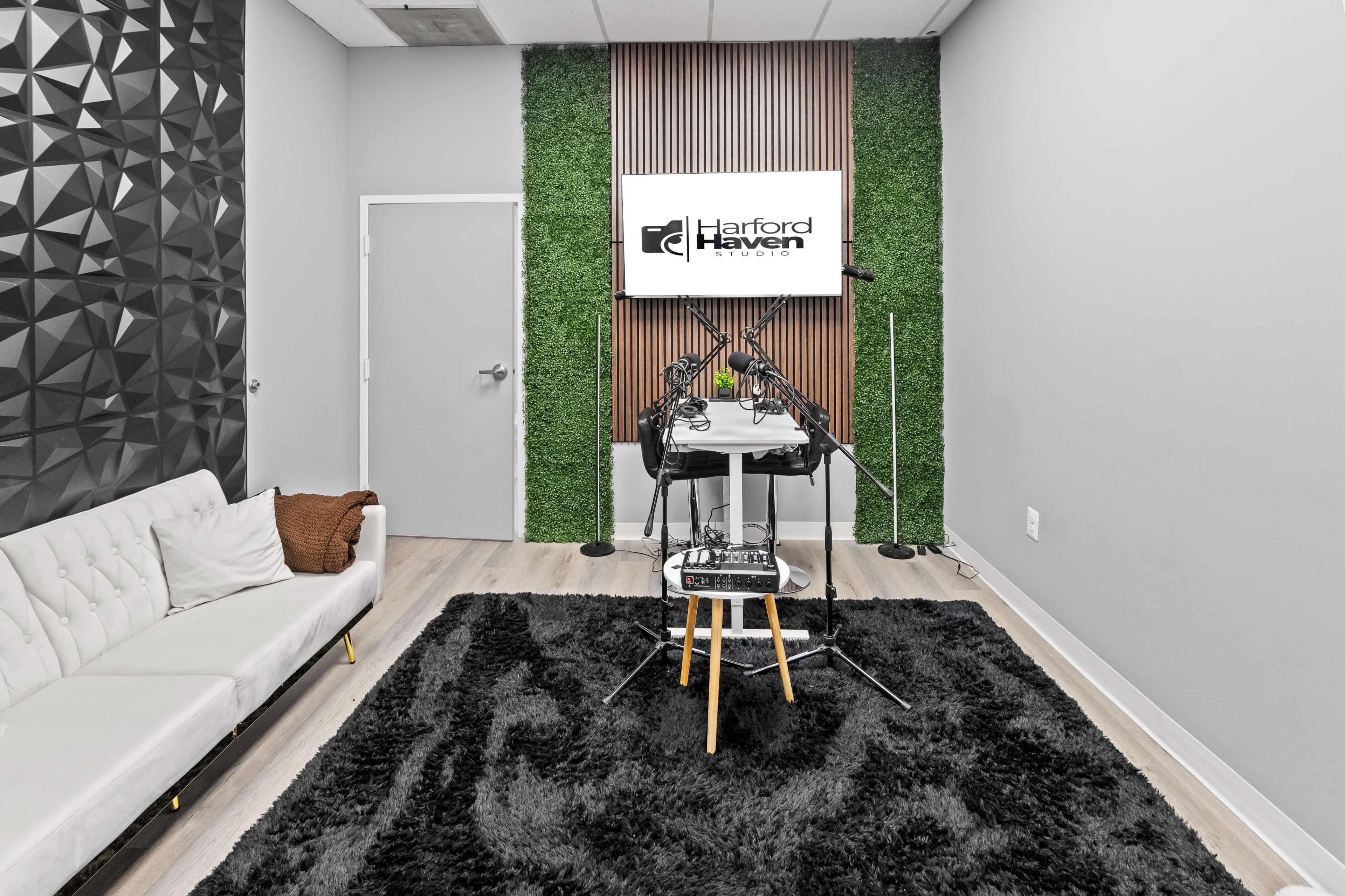 A modern room with a black geometric wall, a green plant wall feature, a logo display, and a setup for audio recording with microphones on a table.