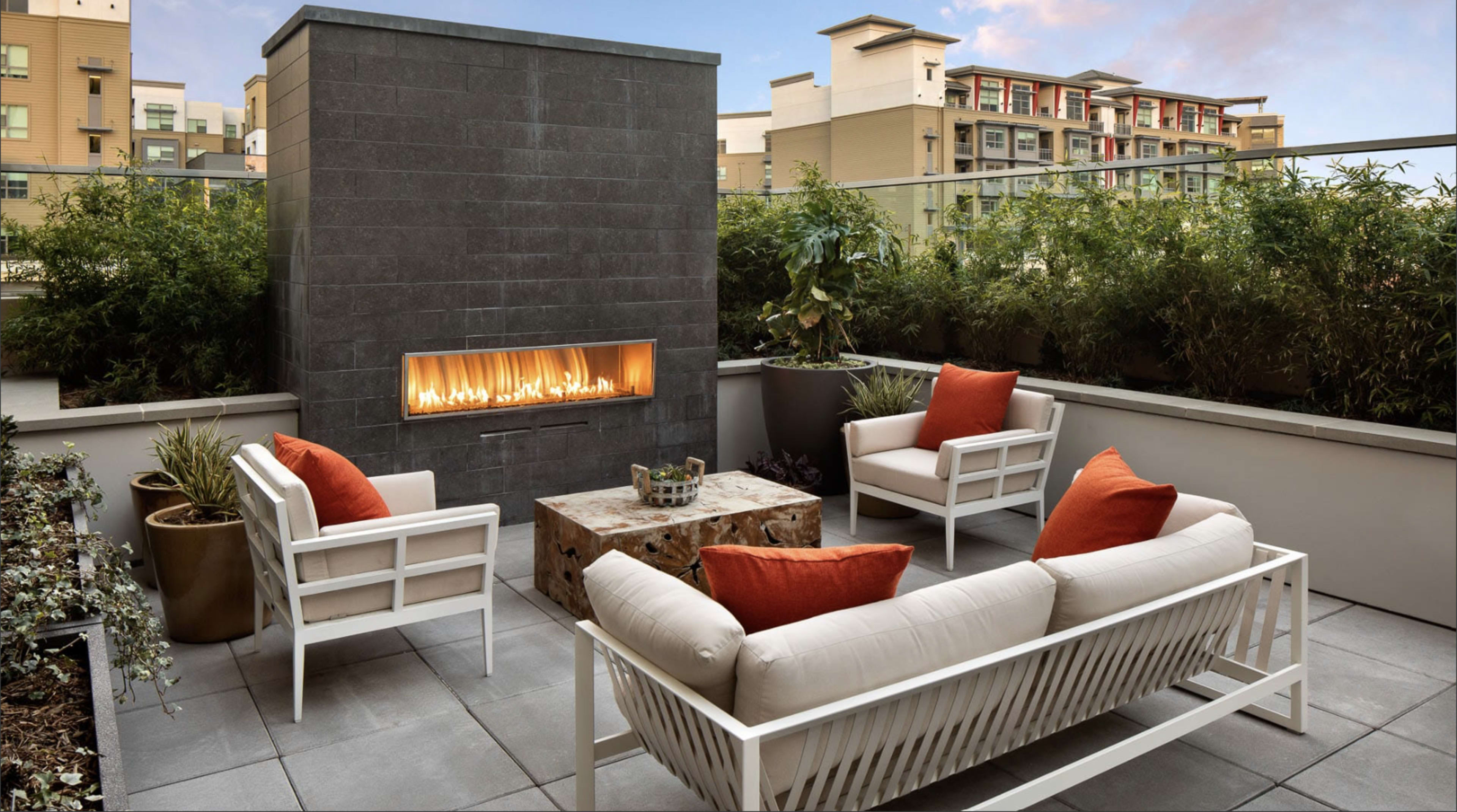 The image shows a modern rooftop patio with a gas fireplace, white outdoor furniture, and potted plants, overlooking a cityscape.