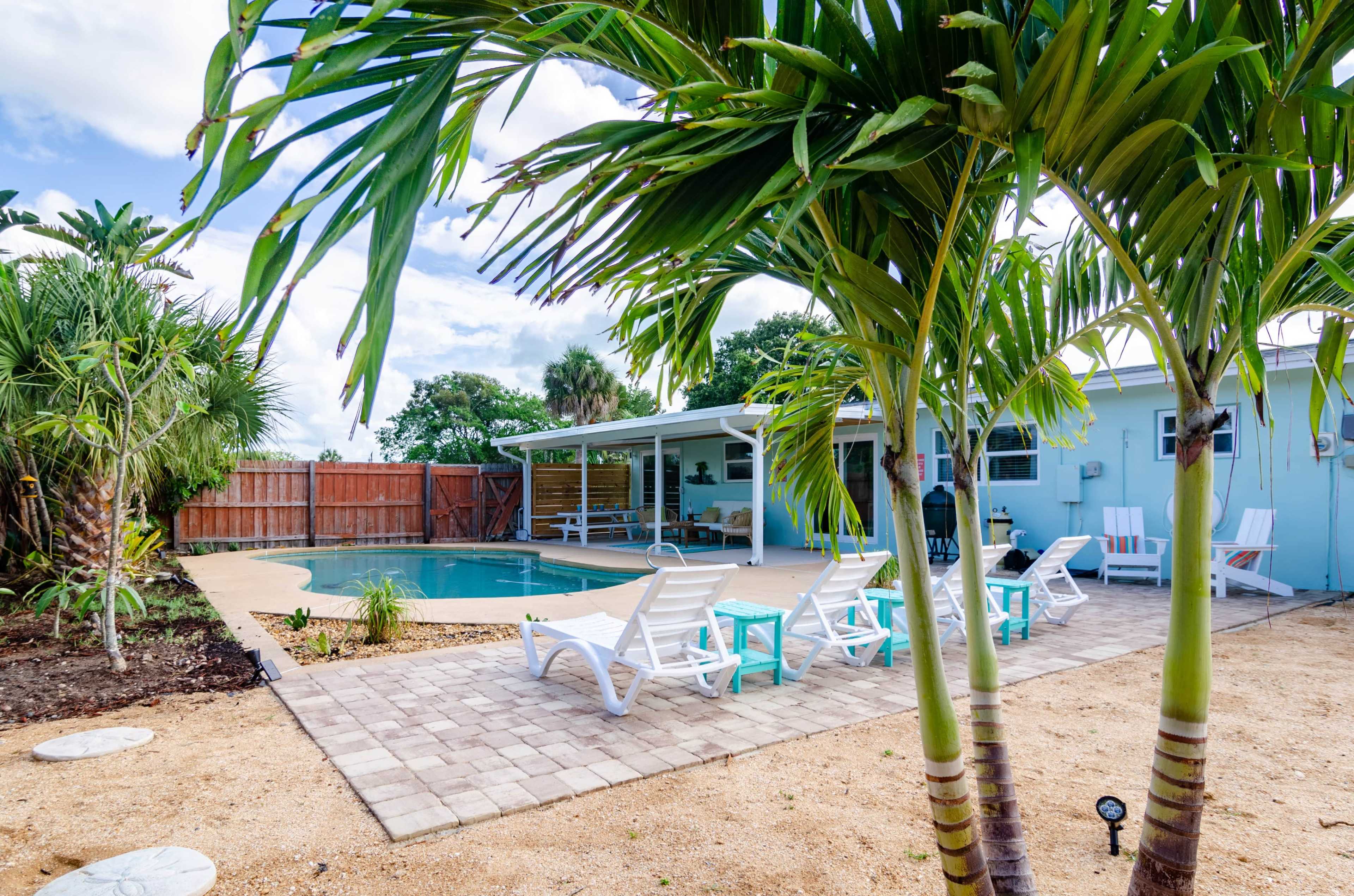 A pool area surrounded by palm trees and lounge chairs, featuring a patio with pavers and a house in the background.