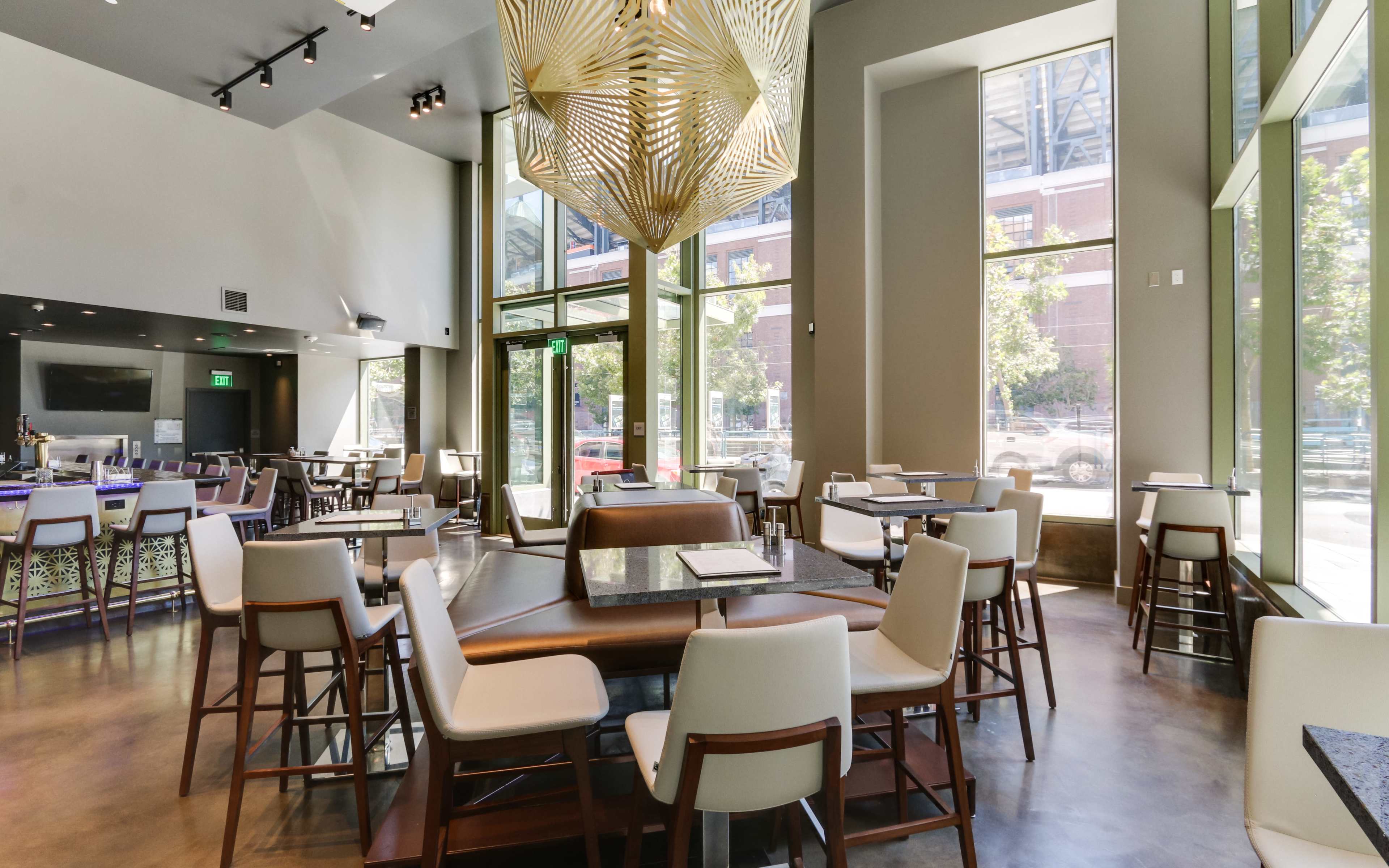 A modern restaurant interior featuring tables and chairs arranged neatly around a large, geometric light fixture, with large windows allowing natural light to enter.