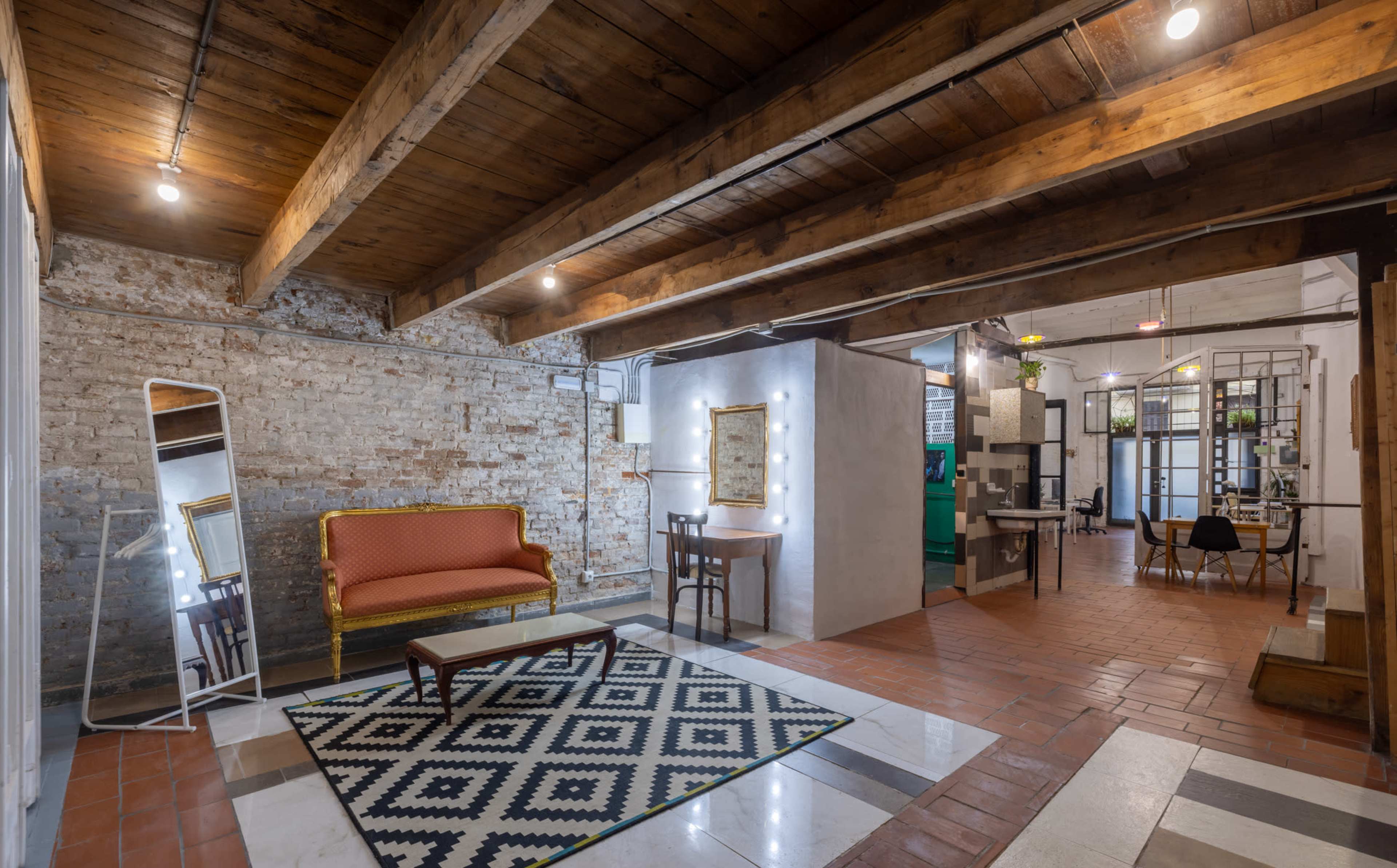 The image shows an interior space featuring exposed wooden beams, a brick wall, a patterned rug, a vintage sofa, and different seating areas separated by partitions.