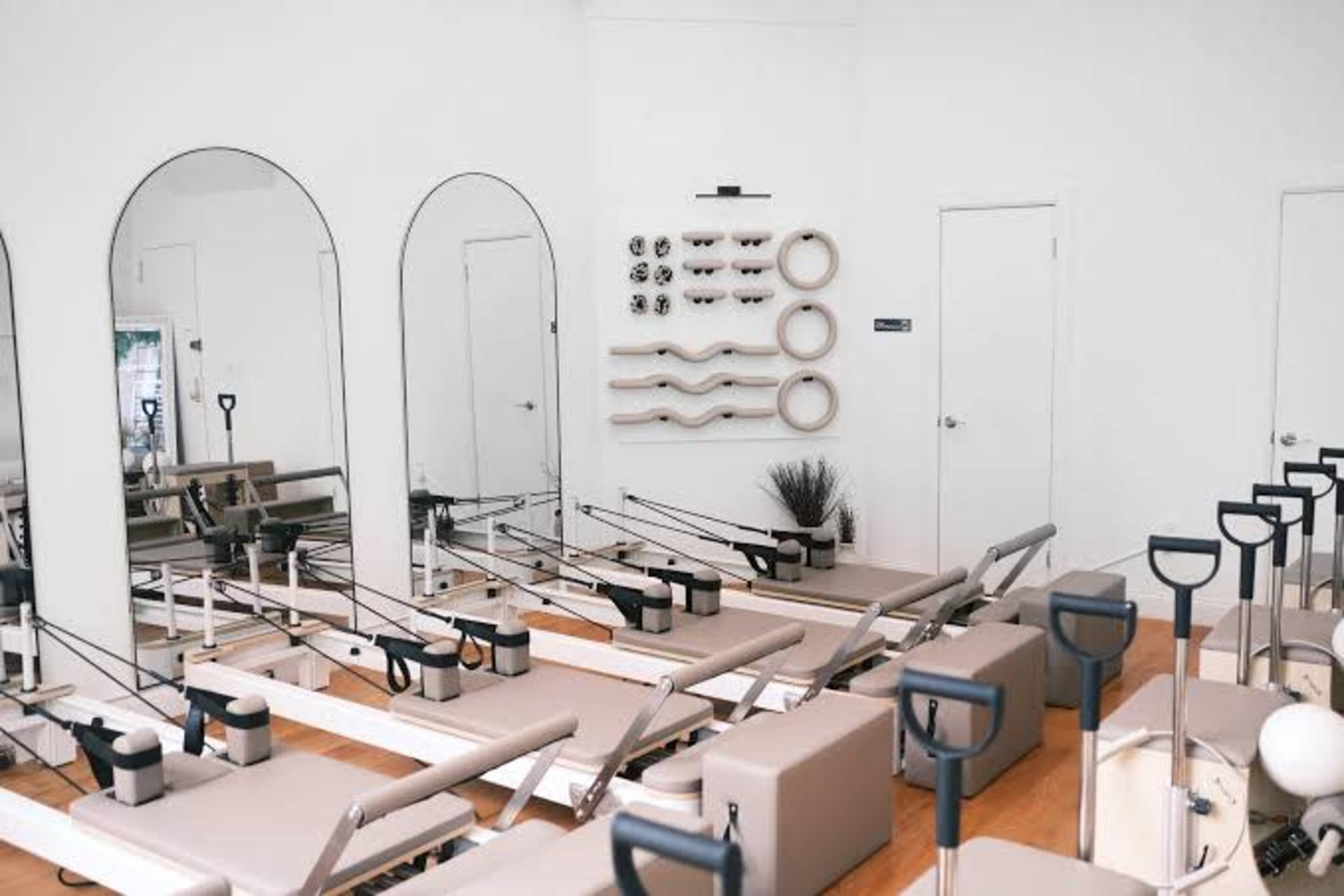 The image depicts a modern pilates studio featuring several reformer machines arranged in a bright, minimalist space with mirrors and various equipment displayed on a wall.
