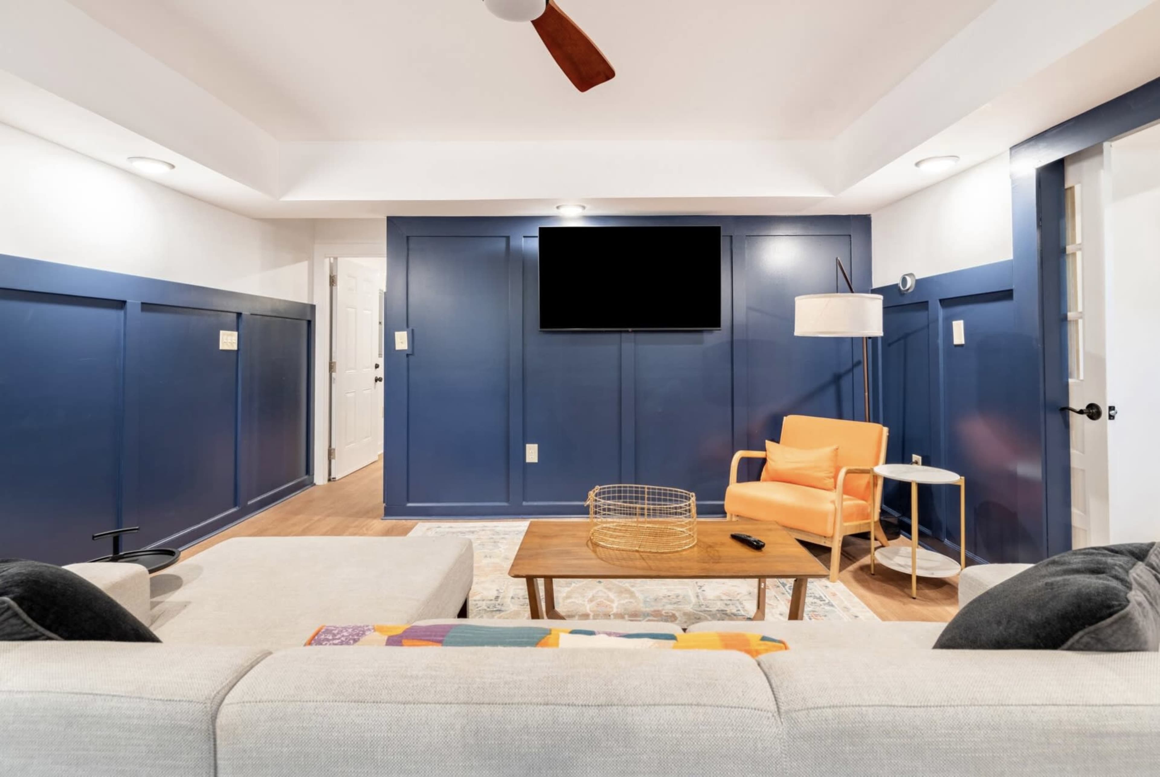 The room features a sofa, a bright orange chair, a wooden coffee table, and a wall-mounted TV, all set against blue-paneled walls and wooden flooring.