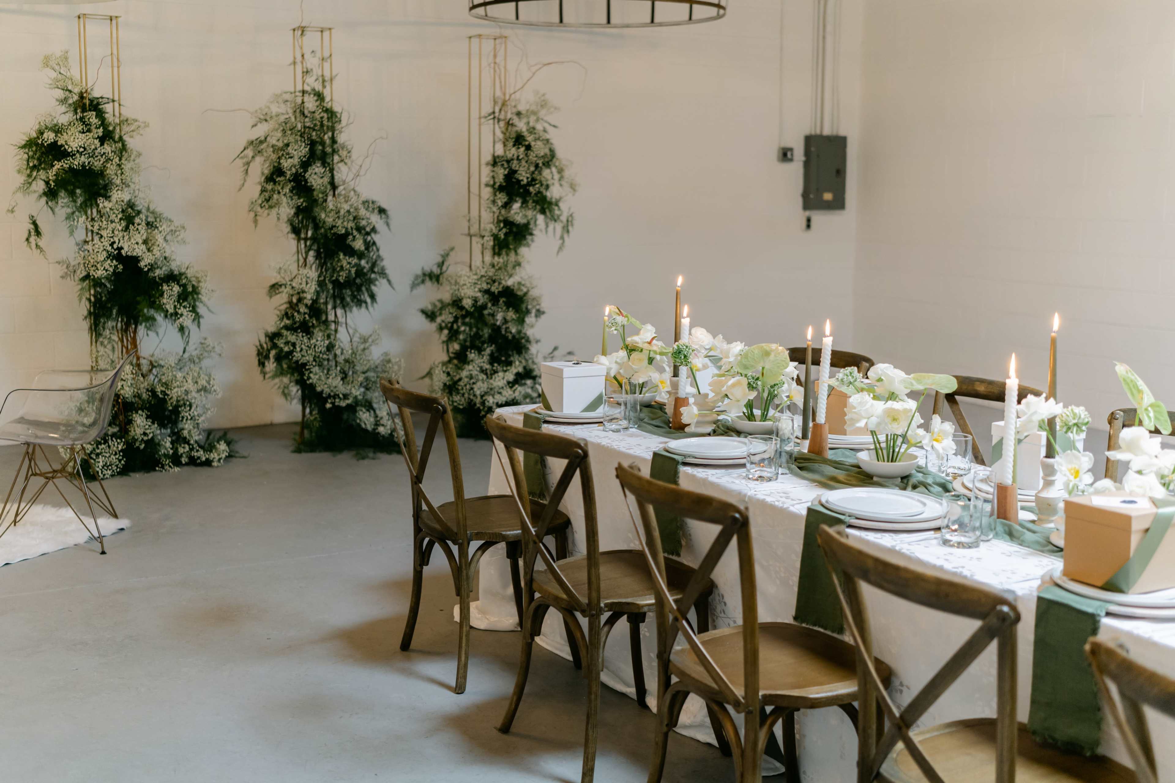 A dining table is elegantly set with floral arrangements and candles, surrounded by wooden chairs, in a minimalist indoor space.