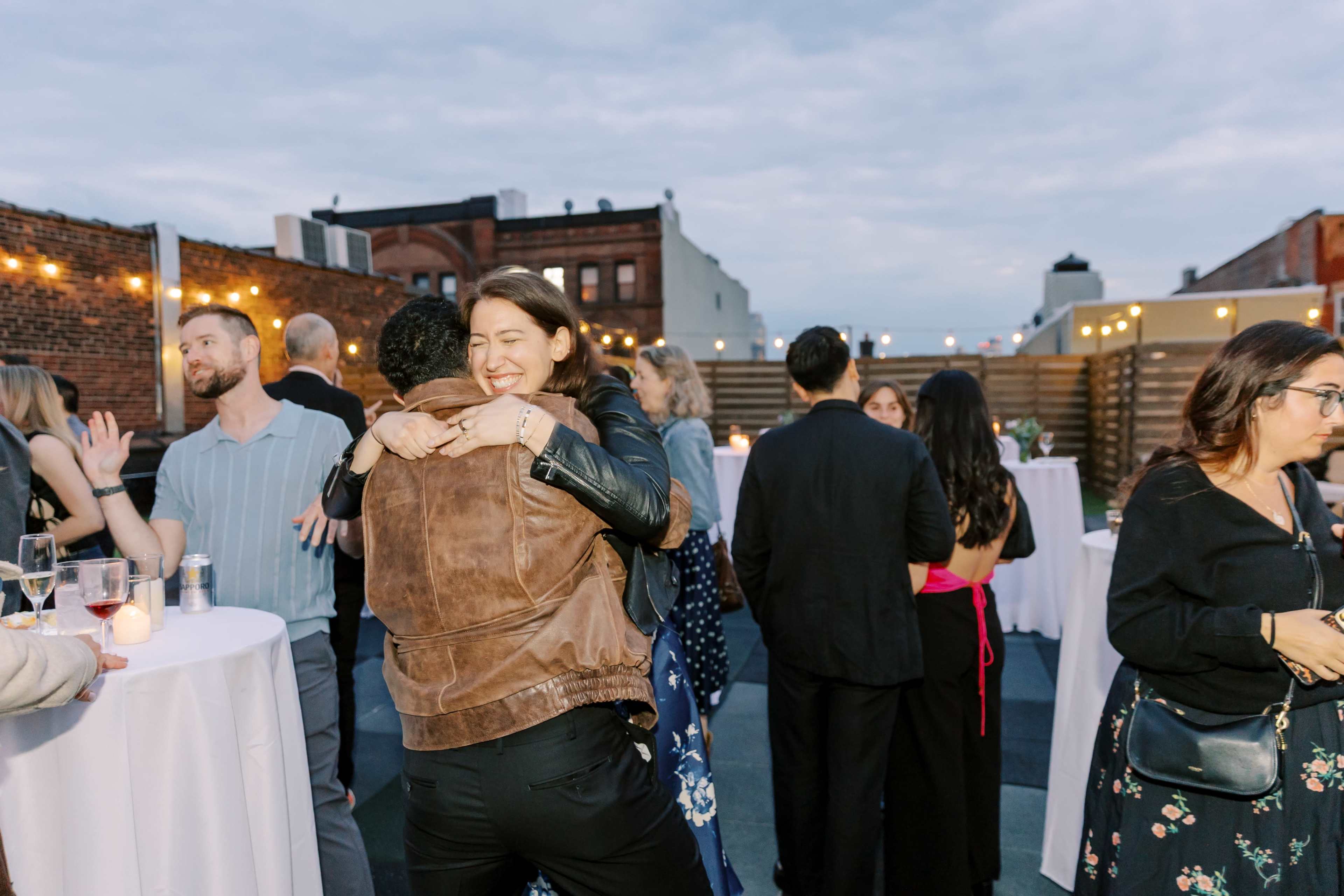 A group of people mingles on a rooftop, with some embracing while others stand at high tables.