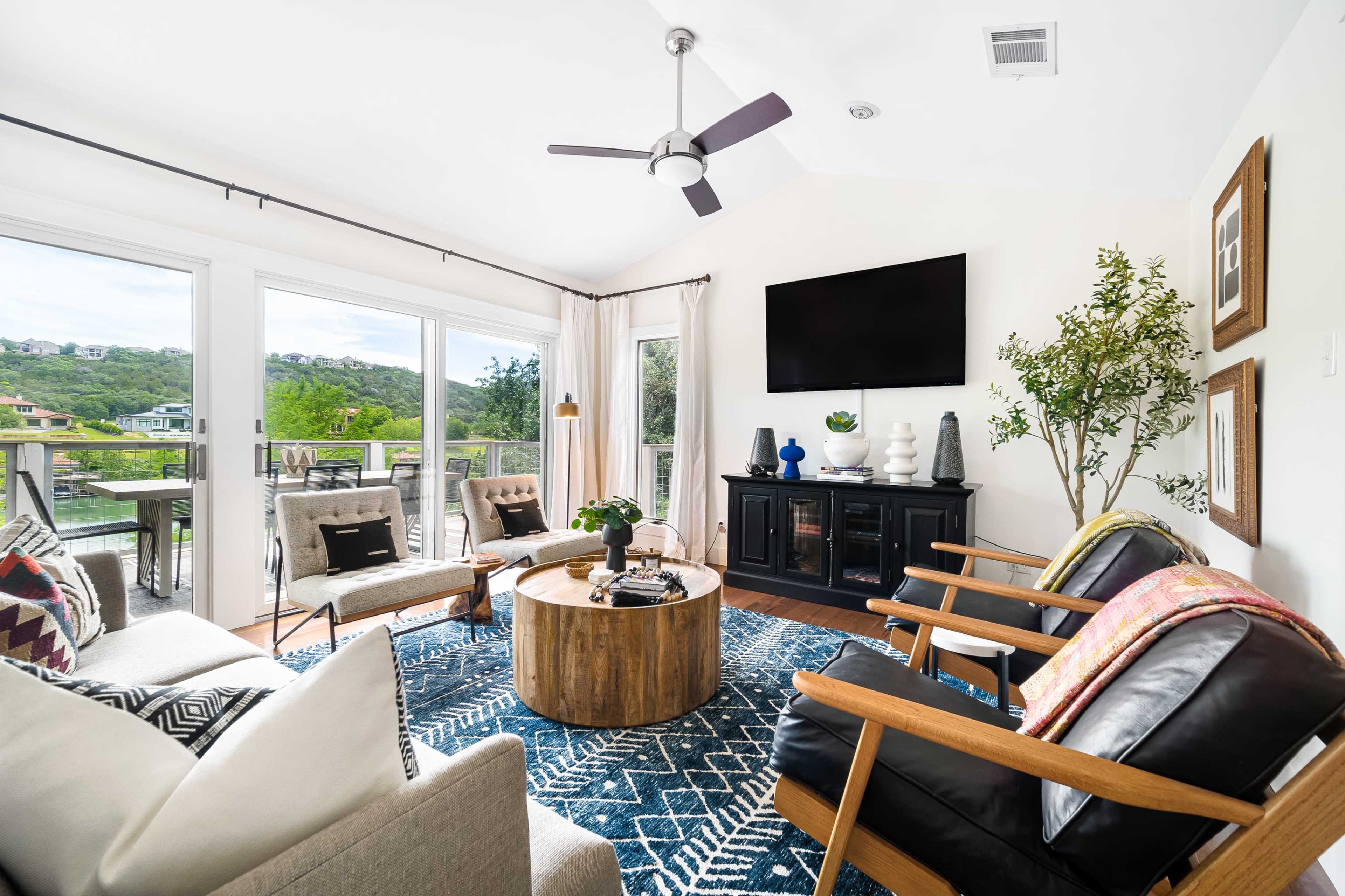 A modern living room with large windows, a round wooden coffee table, and a black cabinet, all arranged around a blue patterned rug.