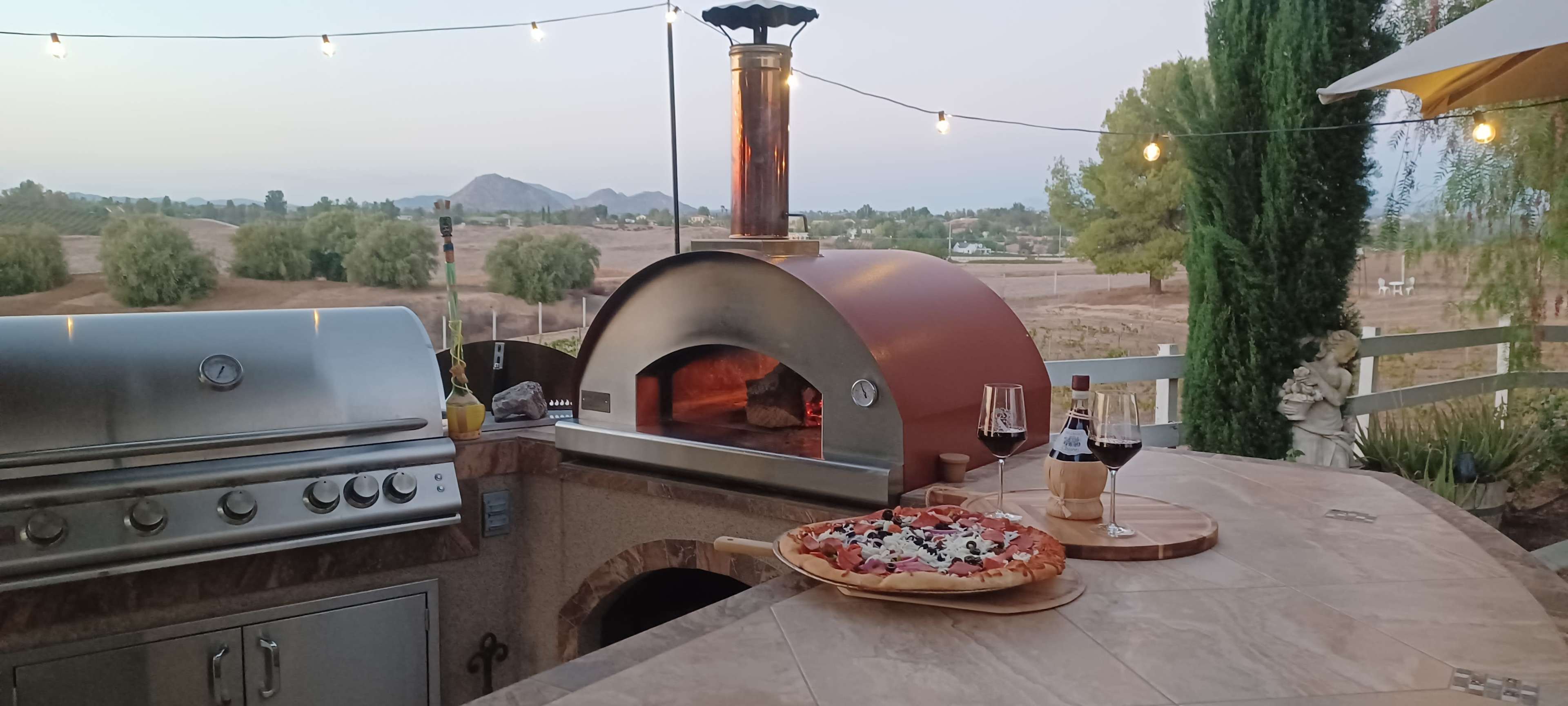 An outdoor kitchen features a pizza oven and grill, with a wooden pizza topped with various ingredients and two glasses of red wine on the counter, set against a scenic backdrop of hills at dusk.