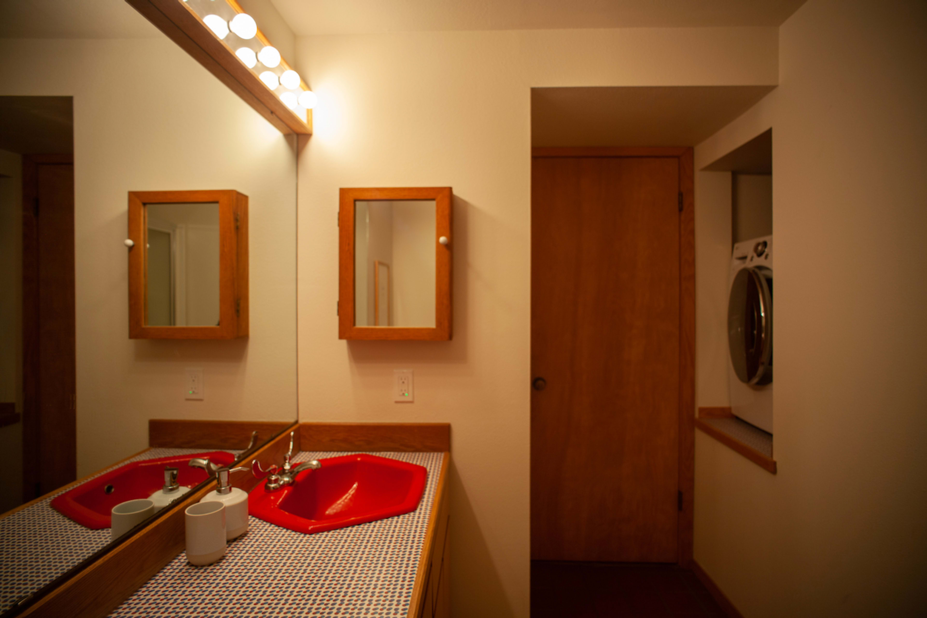 A bathroom with a red sink, a lighted mirror, wooden cabinets, and a door that leads to another room with a stacked washer and dryer.