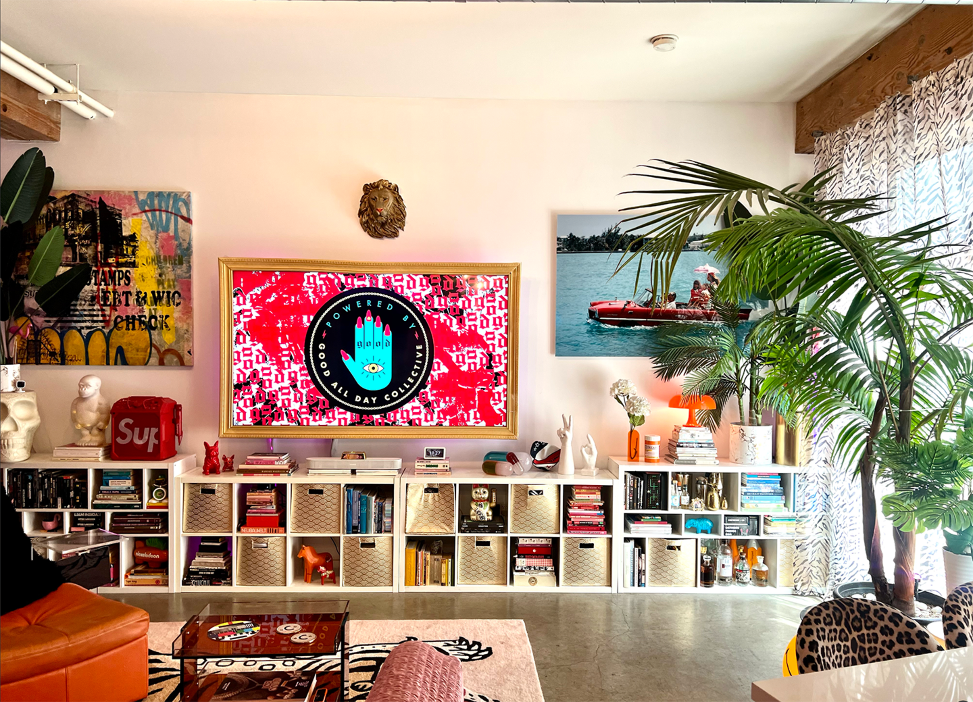 The image shows a modern living room with a large television mounted on the wall, surrounded by bookshelves filled with books and decor, a vibrant plant, and an orange sofa.