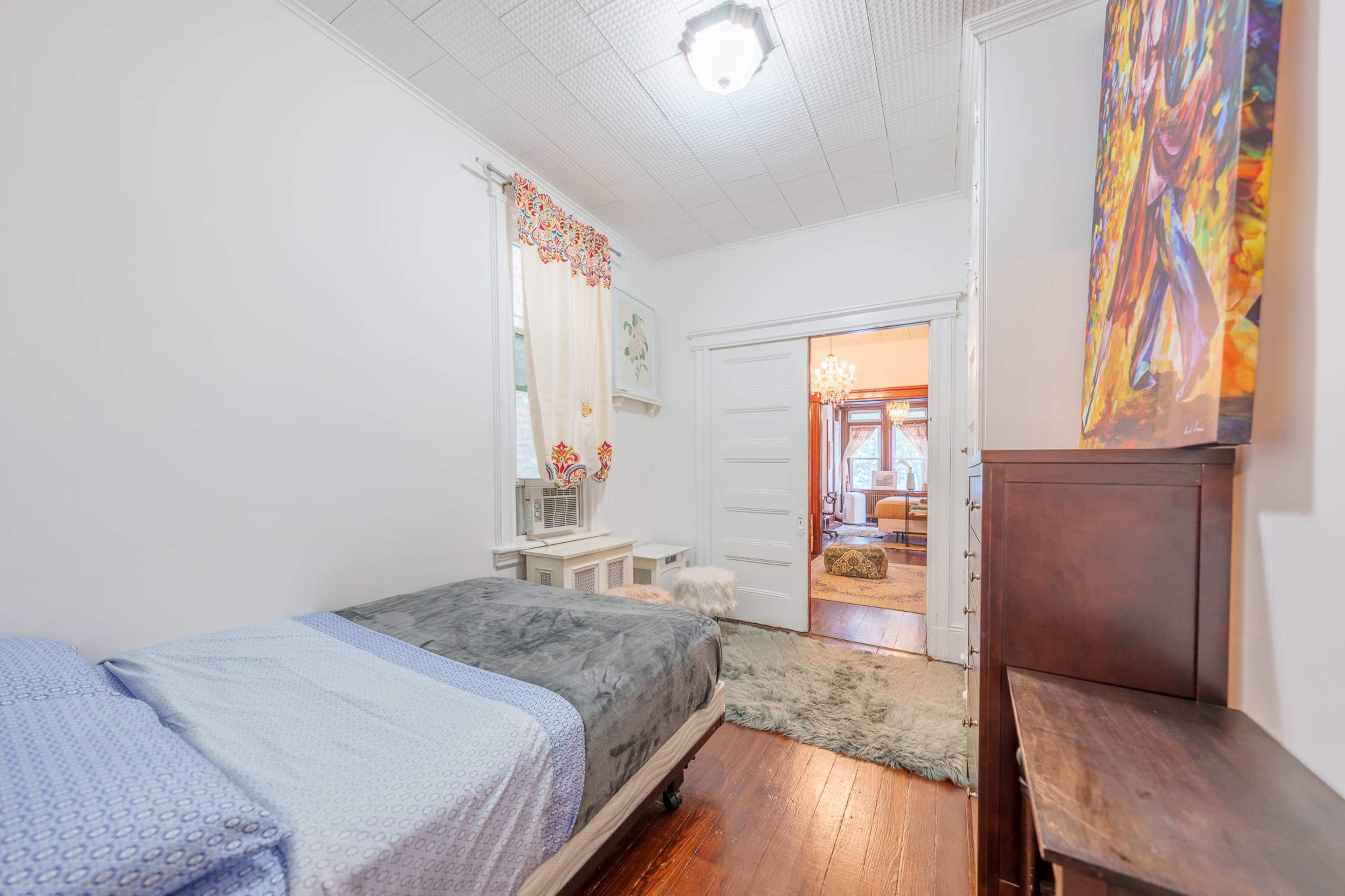 The image shows a small bedroom featuring a bed with a patterned gray blanket and a doorway leading to a living area with ornate decor.