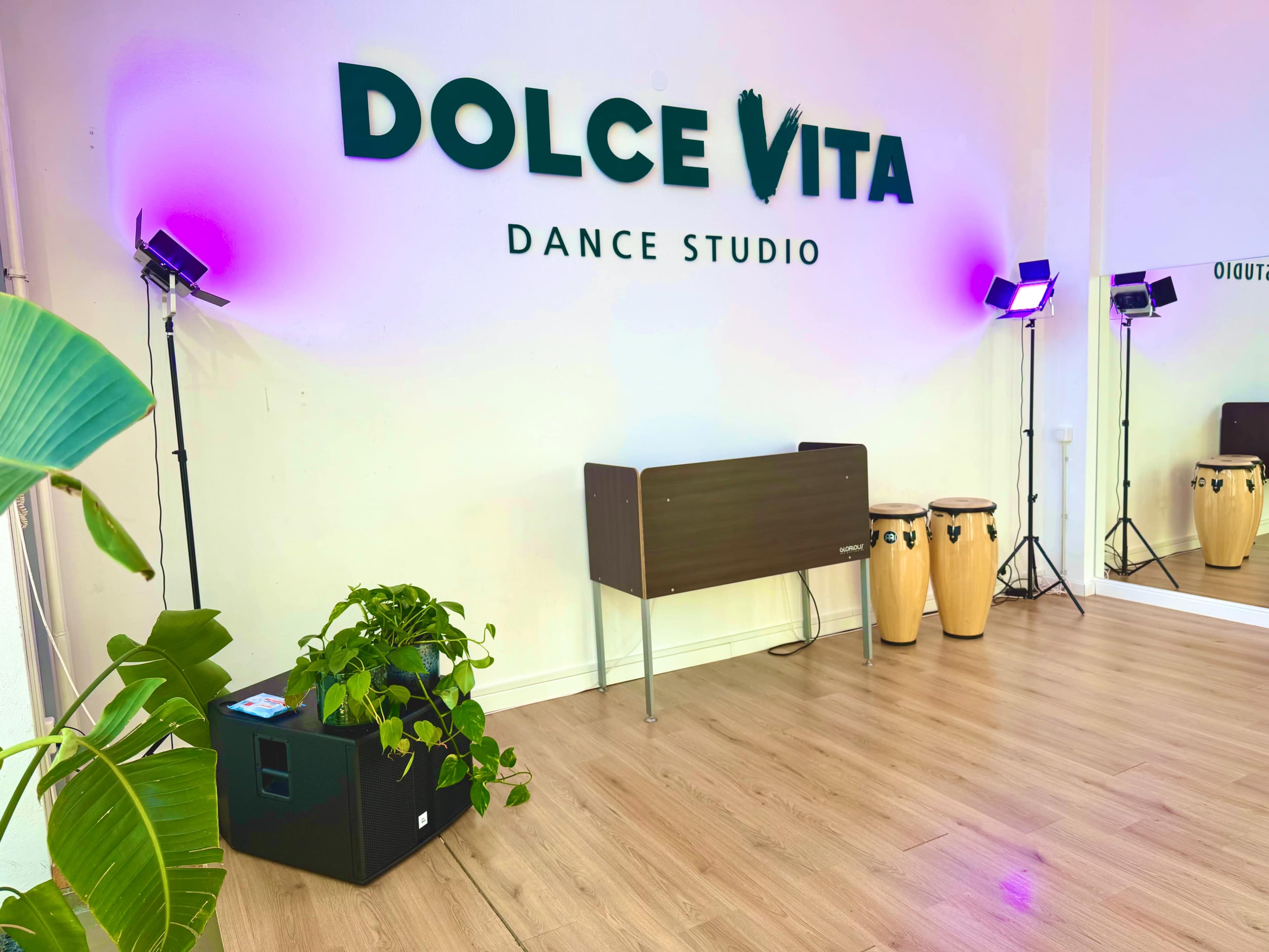 The image shows a dance studio named "Dolce Vita," featuring wooden floors, a plant in the foreground, a conga drum set, and mirrored walls with purple lighting.