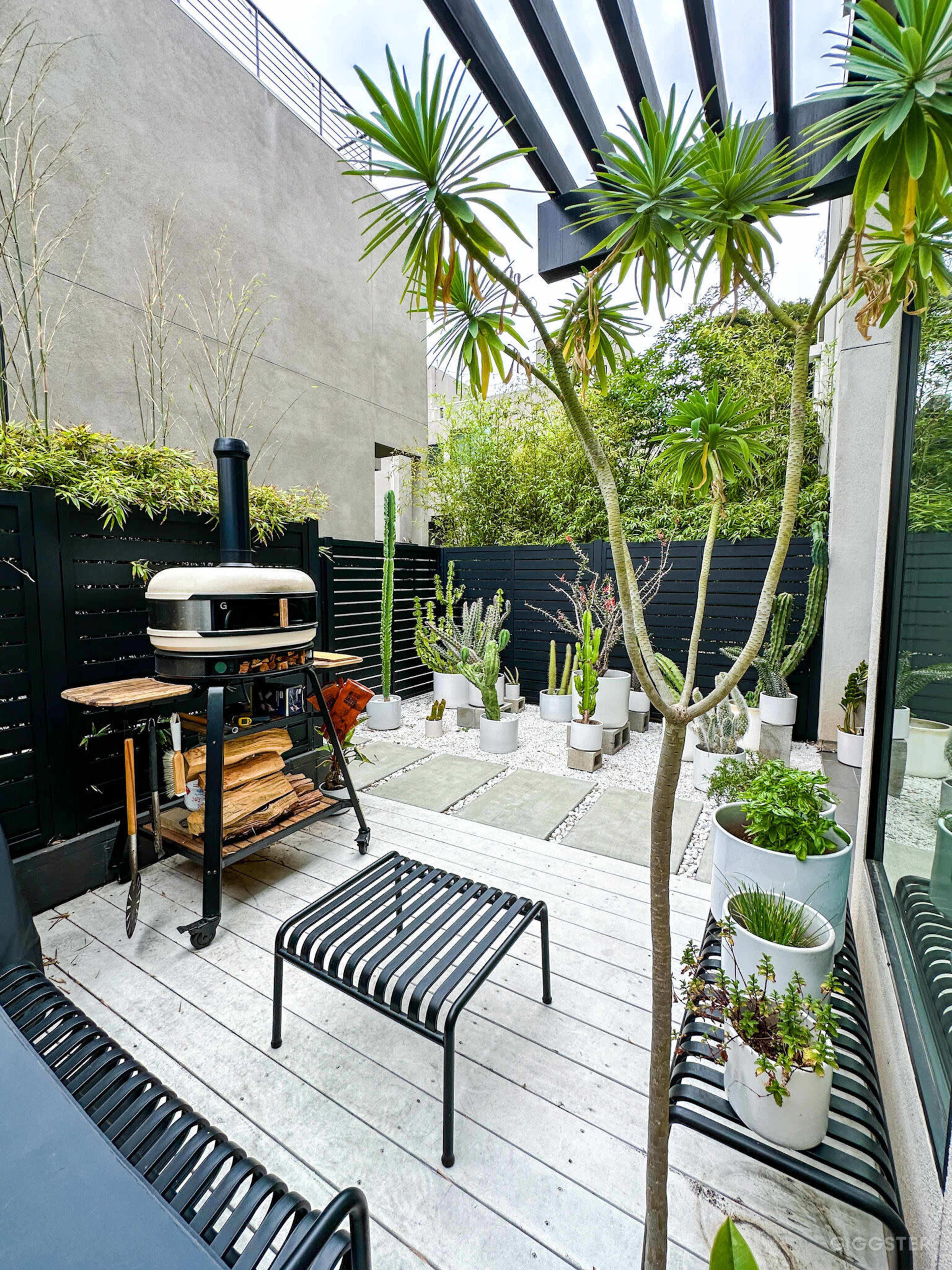 A small outdoor patio with a modern grill, various potted plants, and a combination of stone and wooden pathways.