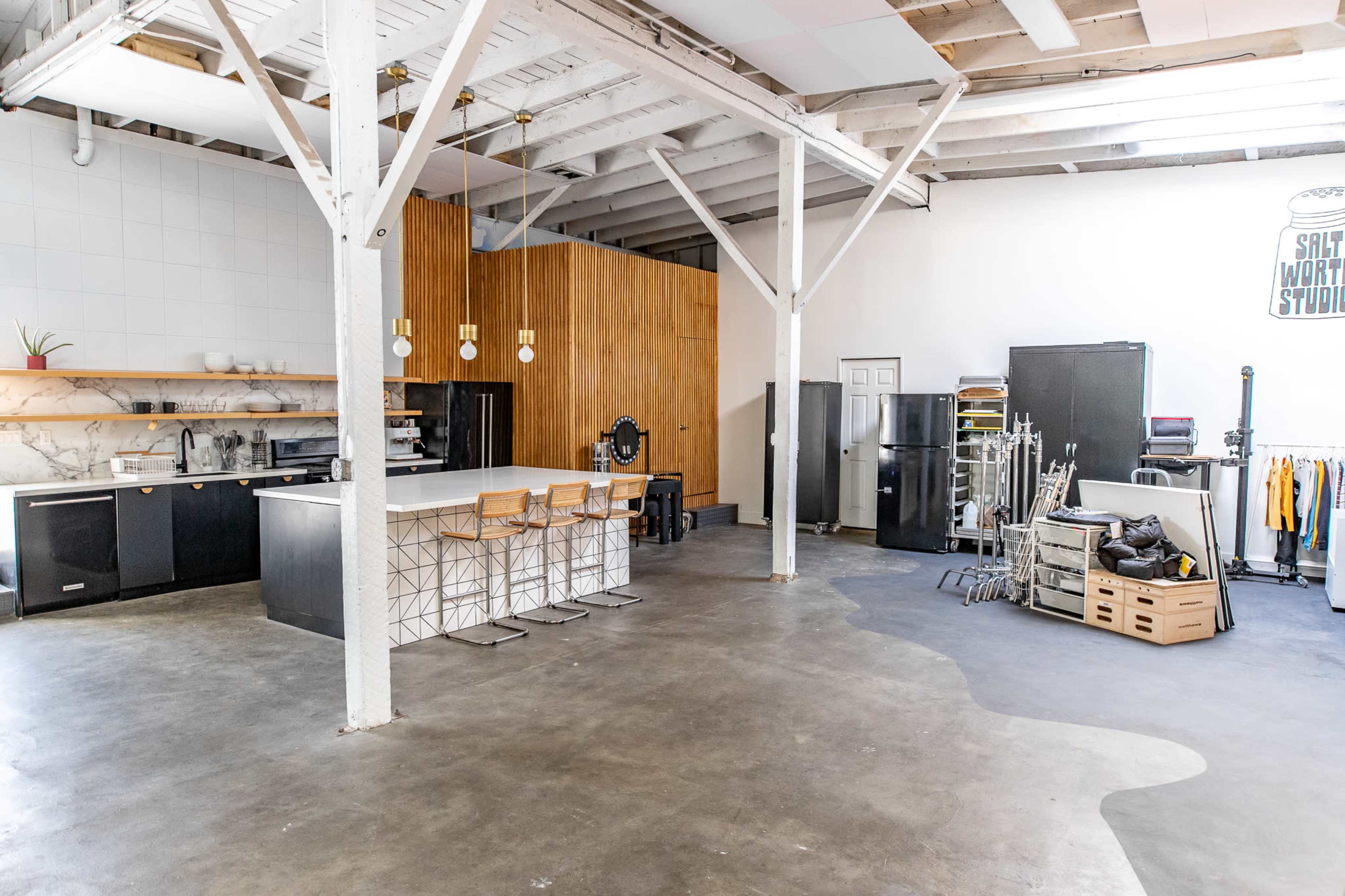 A spacious kitchen area with a large island, black cabinetry, and seating, alongside a cluttered workspace filled with various equipment and clothing.