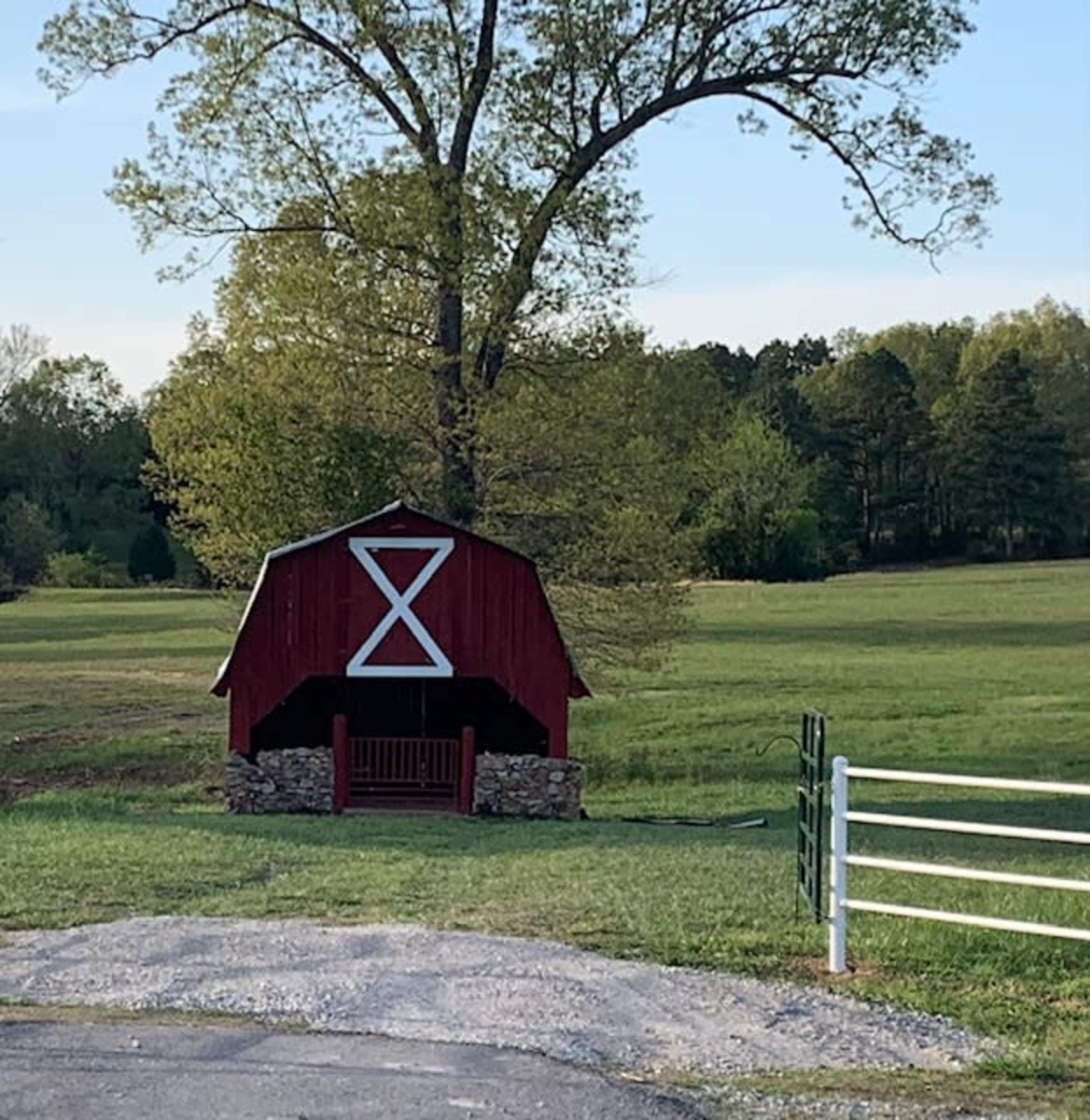 A red barn with a white cross on its door stands in a green field near a tree and a fenced area.