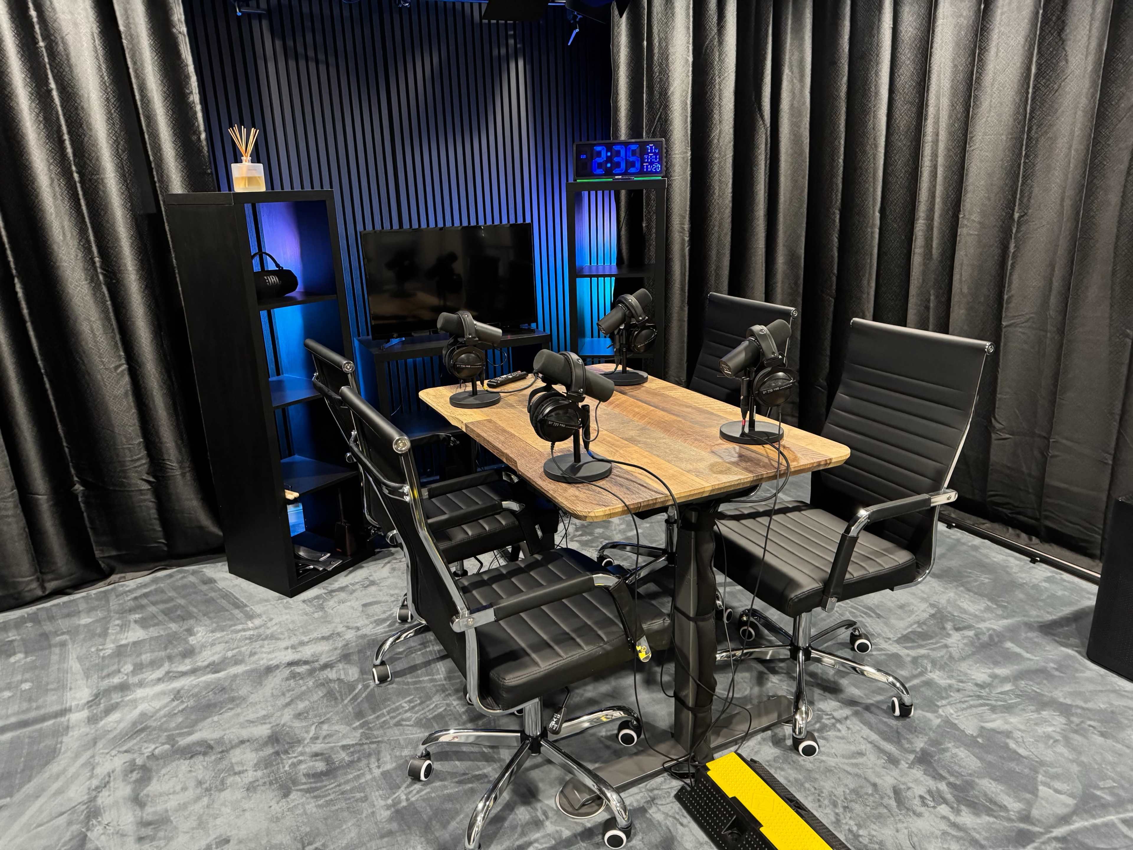 A podcast recording studio features a wooden table with six black leather chairs and multiple microphones arranged for recording.