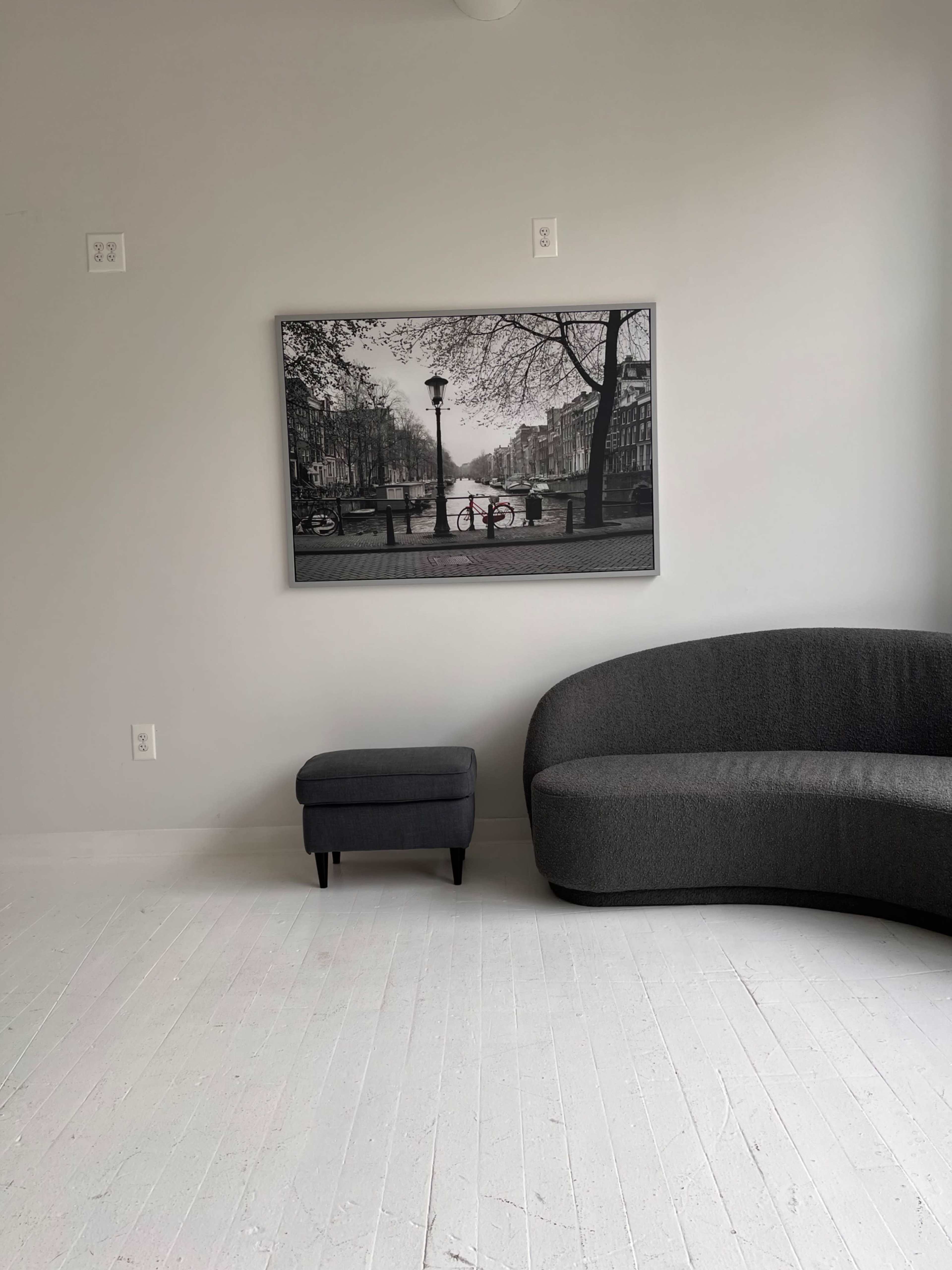A modern, minimalist room with a gray curved sofa, a small gray stool, and a black and white photograph of a canal scene on the wall.