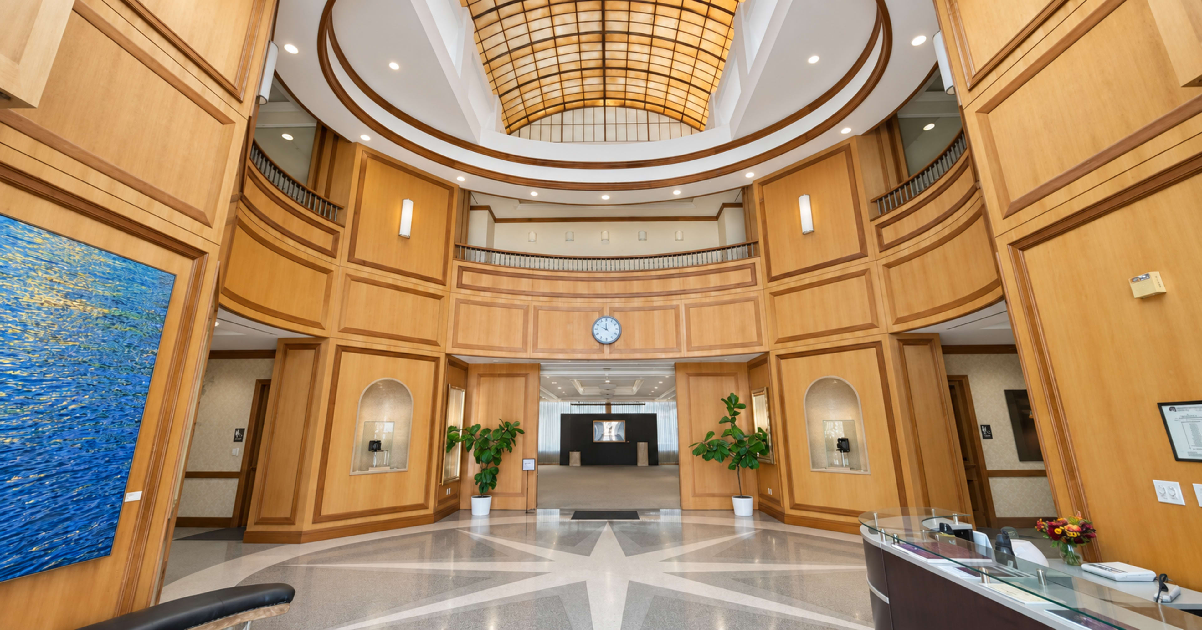 The image shows a modern lobby with a high ceiling, wooden paneling, large plants, and a reception area featuring a curved design.