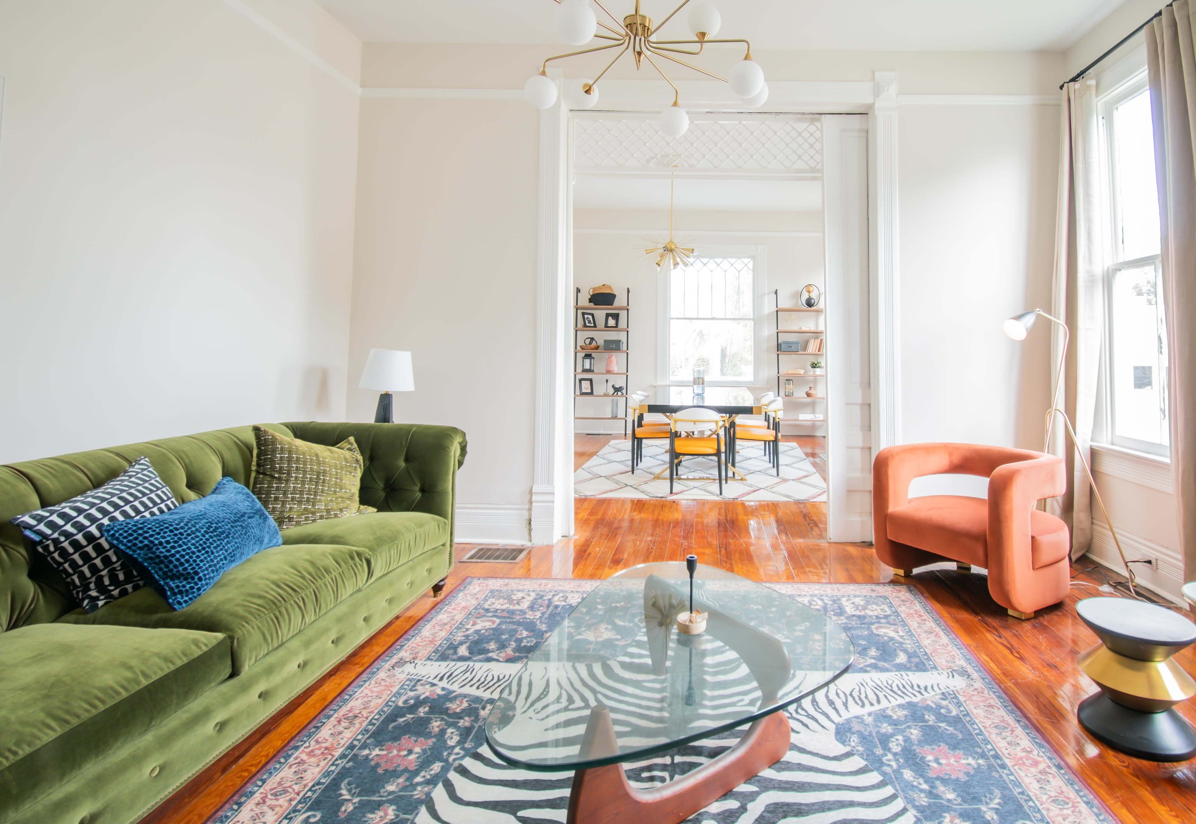A green velvet sofa and an orange armchair are arranged in a bright room with hardwood floors and large windows, featuring a glass coffee table and a black and white zebra-patterned rug.