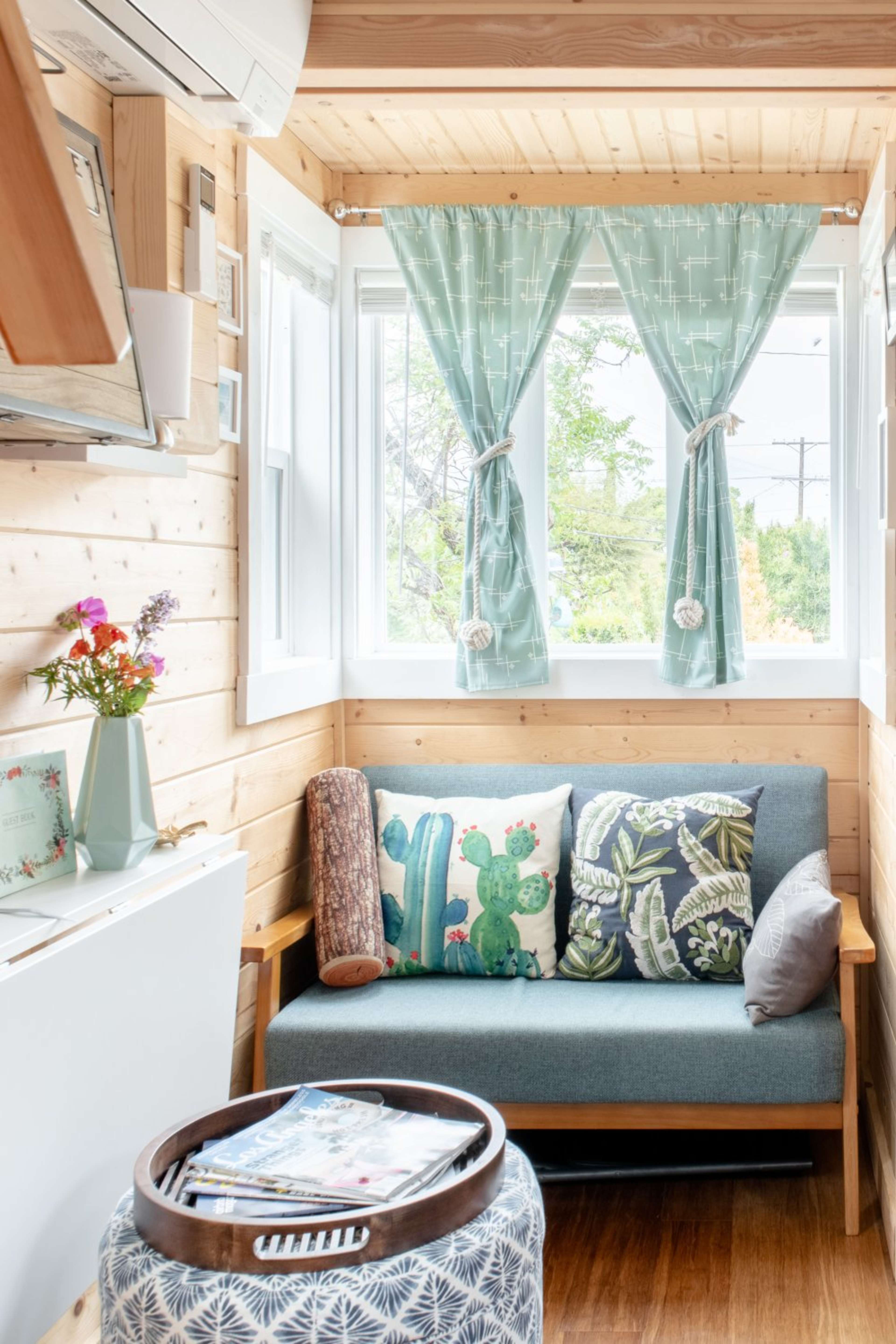 A cozy interior of a wooden room with a sofa adorned with decorative pillows, a coffee table with magazines, and a window with light blue curtains.