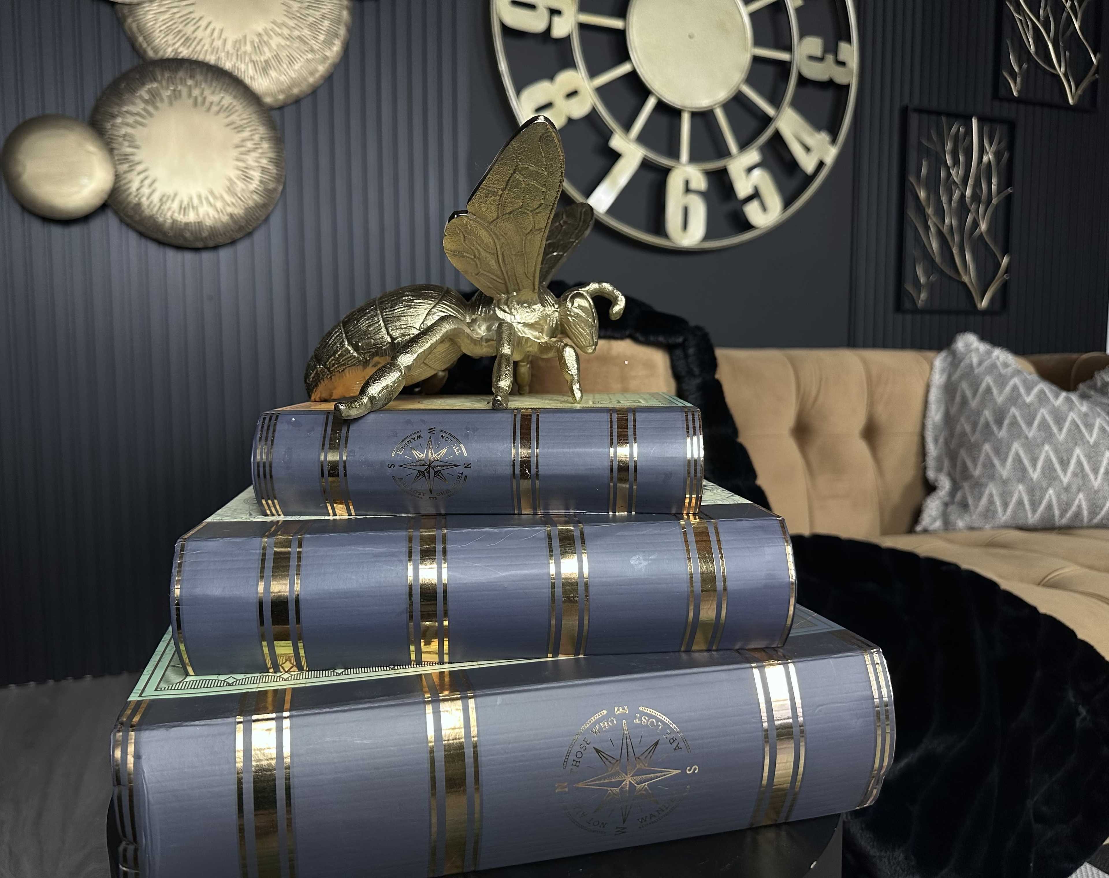 A golden decorative insect is positioned atop a stack of blue books on a table in a living room with a large wall clock and decorative plates.