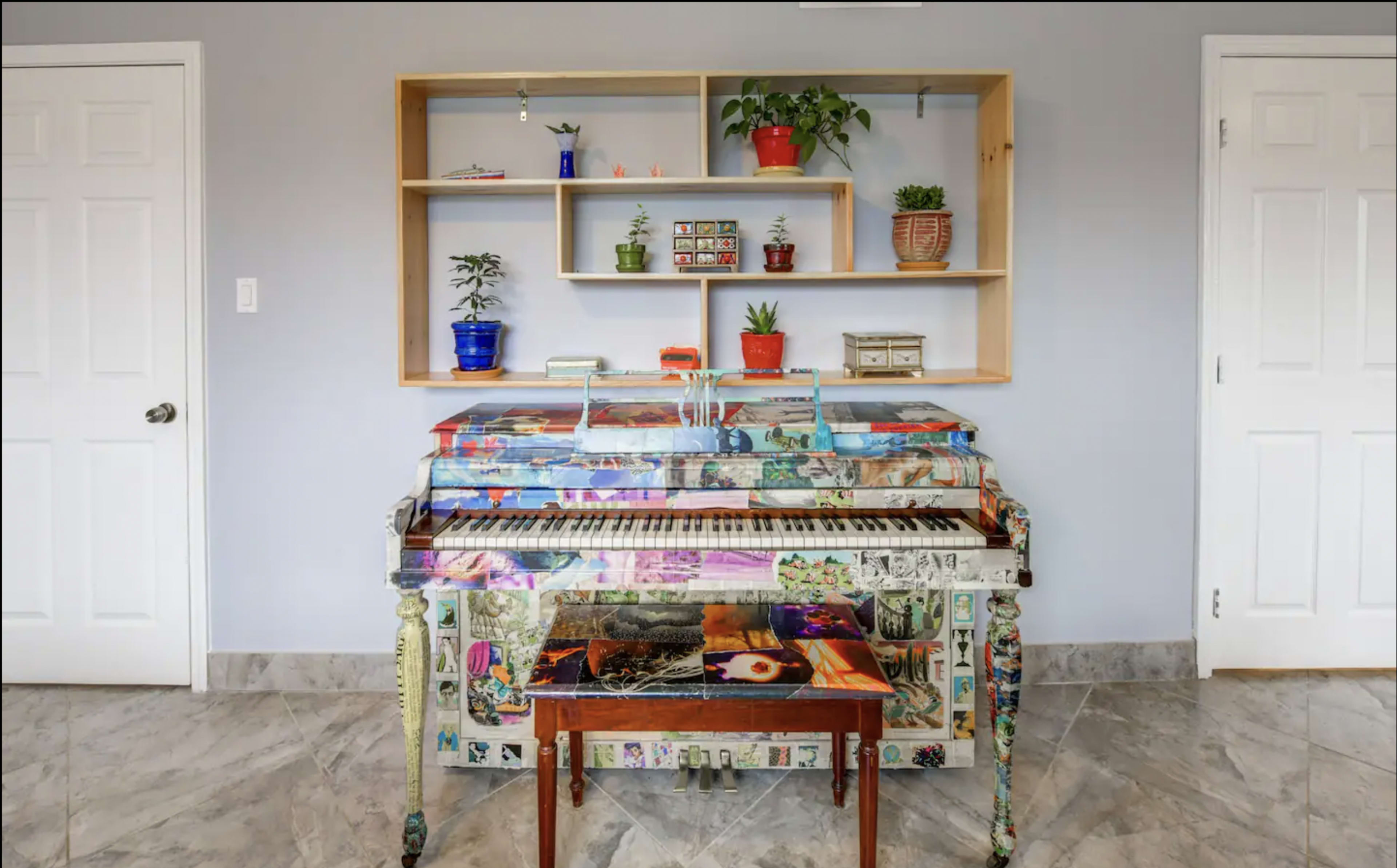 A decorated piano with colorful artwork and a small wooden table sits beneath a shelf containing various plants and decorative items against a plain wall.