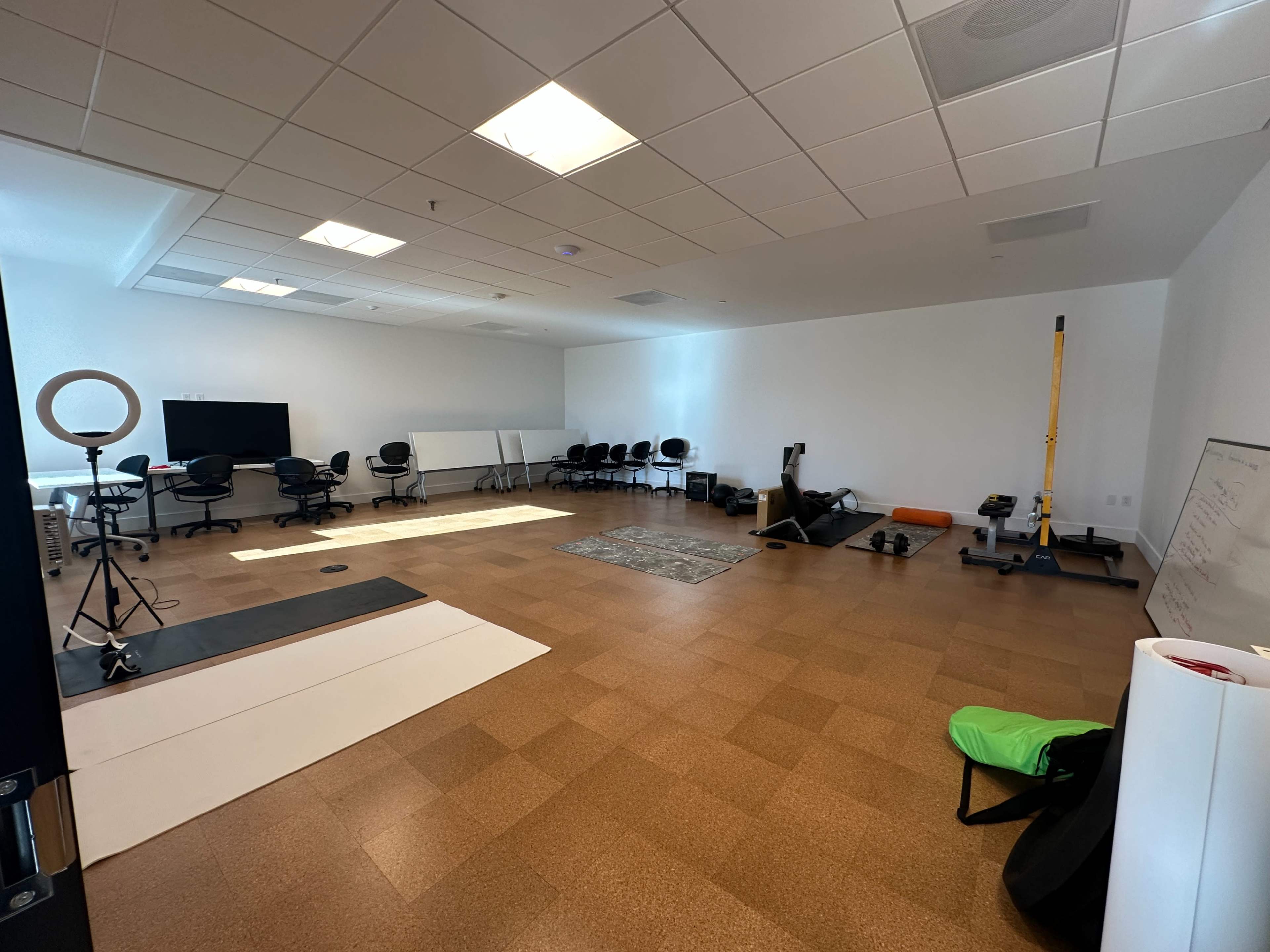The image shows a spacious, empty room with various chairs arranged in a semi-circle, exercise equipment in one corner, and a large screen against one wall.