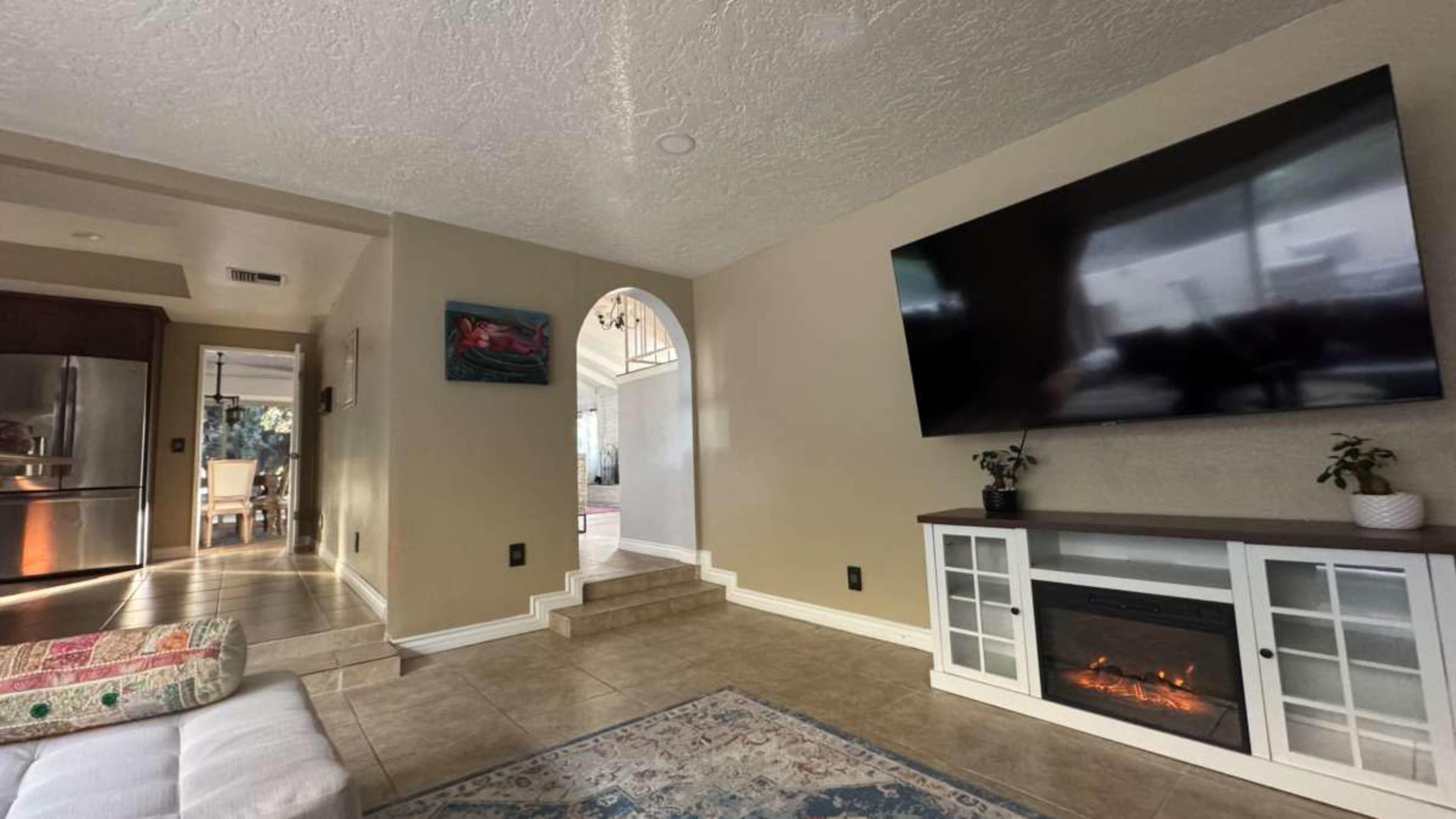 The image shows a living room featuring a mounted television, a faux fireplace unit, and a view into a neighboring room with a kitchen area visible at the back.