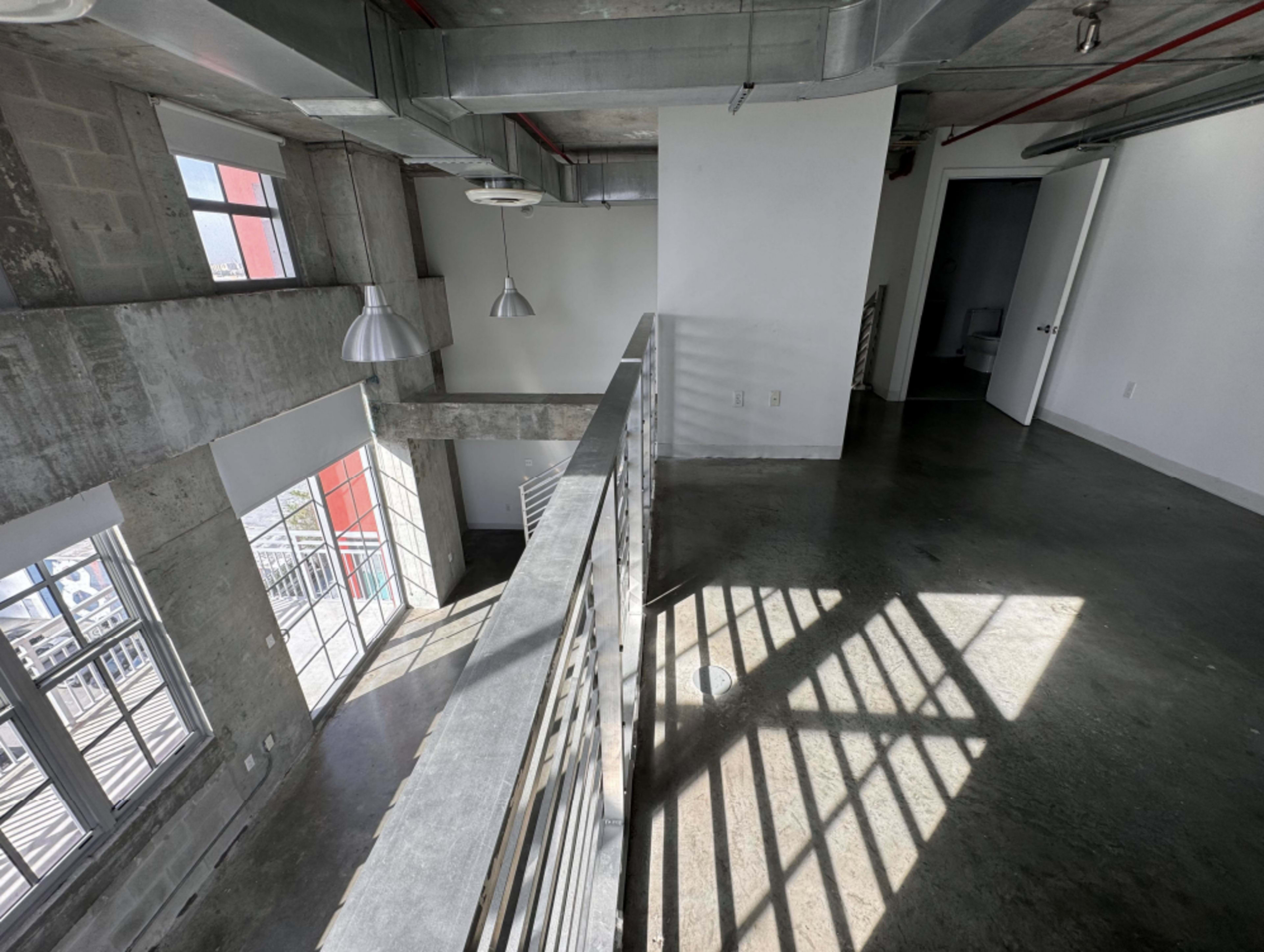 The image shows an industrial-style interior space with concrete walls, metal railings, and large windows that allow natural light to fill the area.