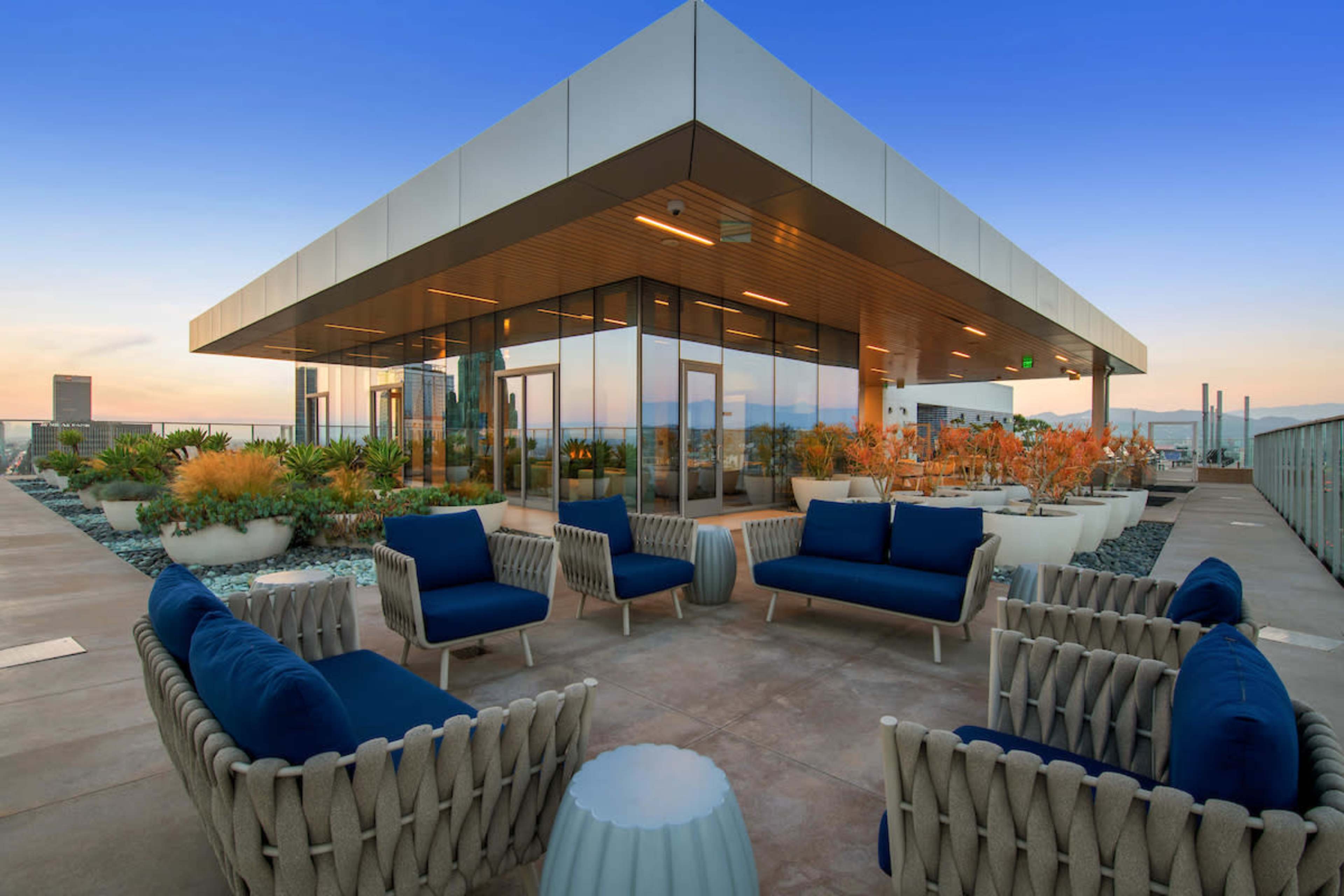 The image shows a modern rooftop lounge area with comfortable seating and planters, surrounded by the skyline at dusk.