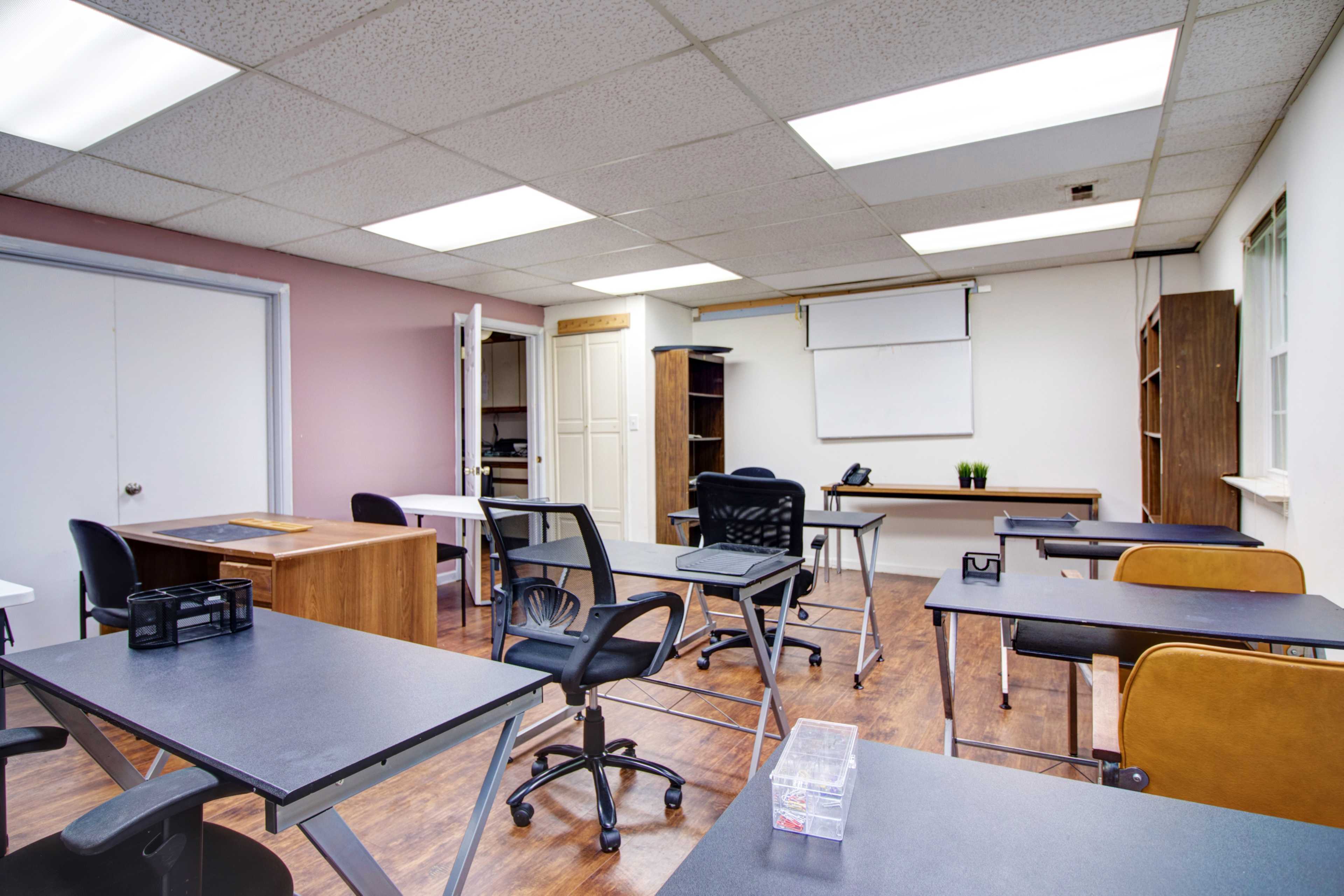 The classroom features several desks and chairs arranged with a whiteboard and shelves along the walls.