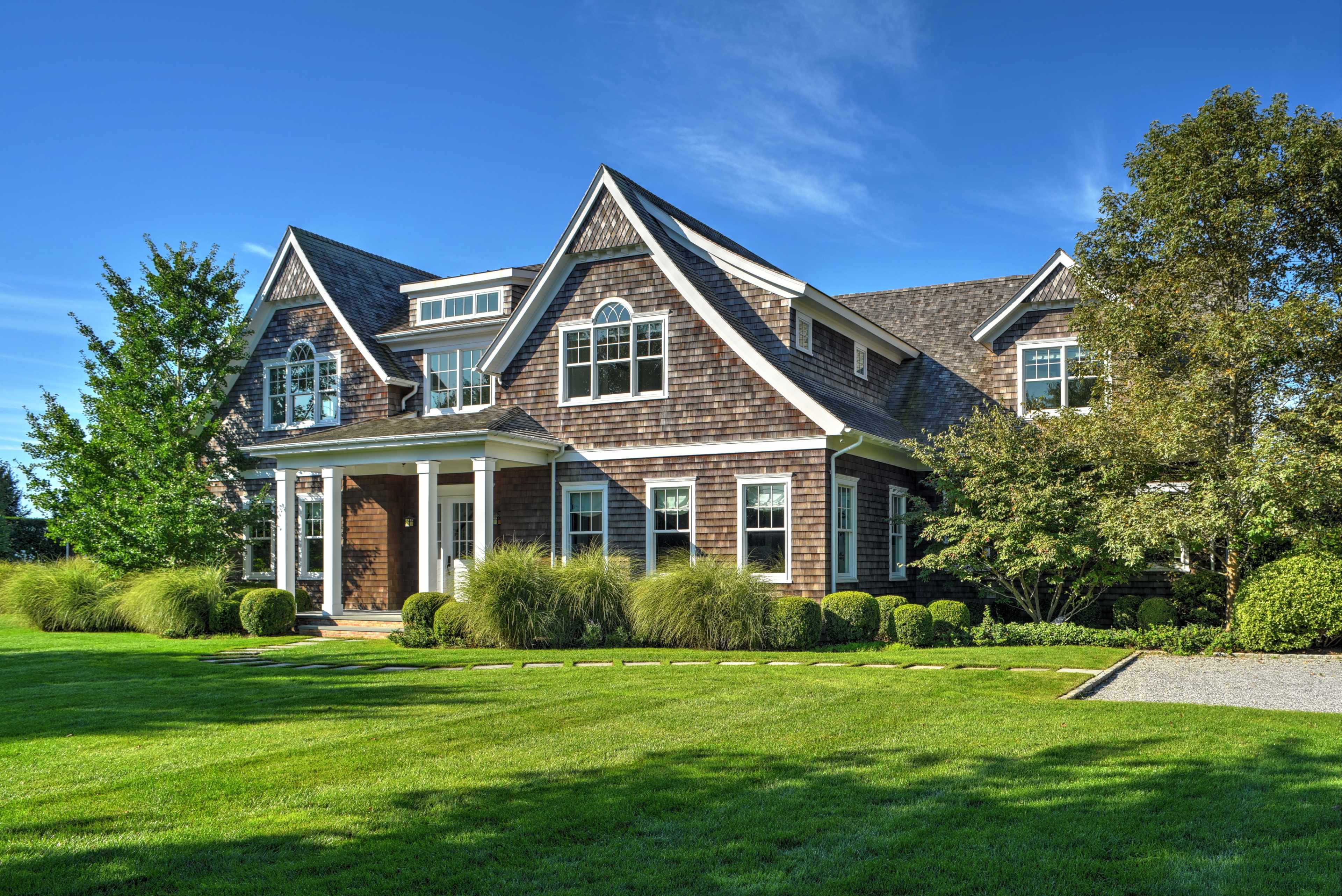 A large, two-story house with a shingled exterior is situated on a manicured lawn surrounded by neatly trimmed bushes and pathways.