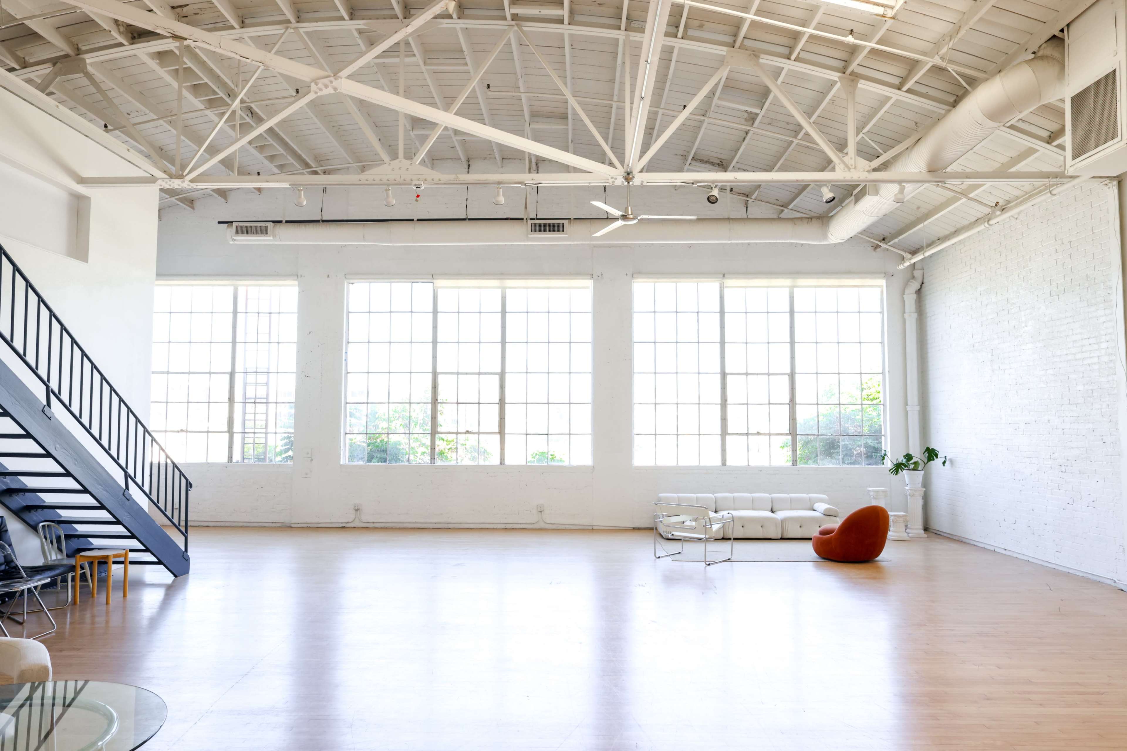 The image shows a spacious, well-lit interior with large windows, a staircase, a white sofa, and a singular orange chair.