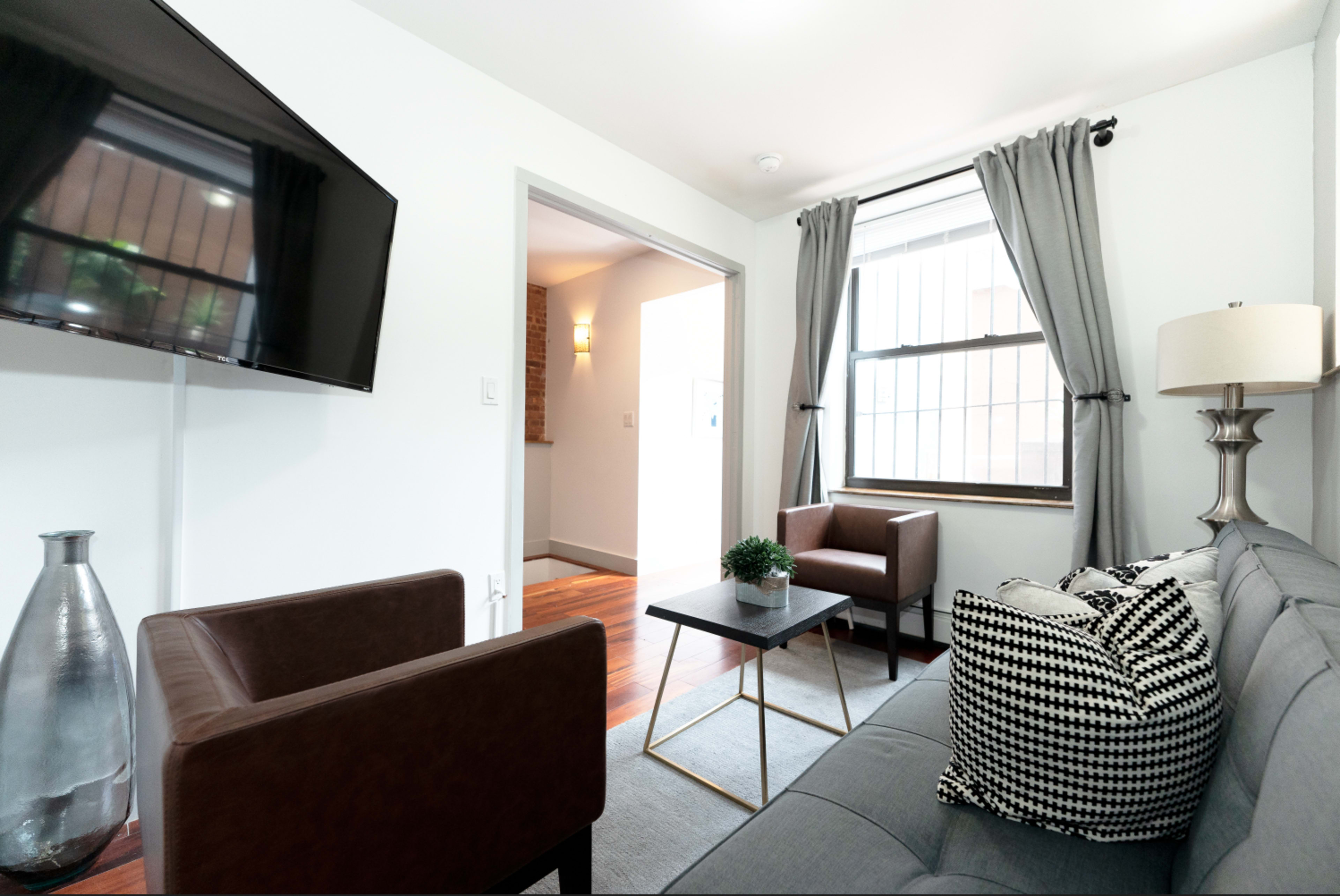 A modern living room features a gray sofa, two brown chairs, a small coffee table, and a television mounted on the wall.
